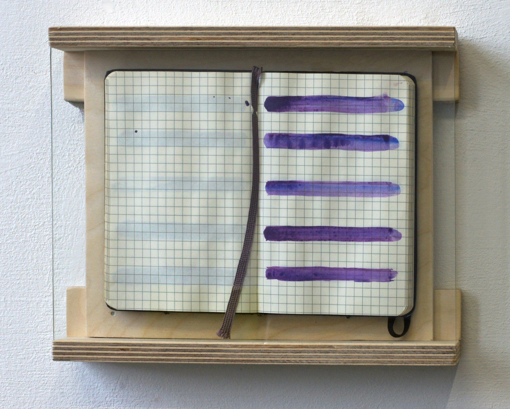 Een open notitieboek met ruitjespapier en paarse horizontale verfstrepen, ingelijst in een houten frame tegen een muur met structuurverf.