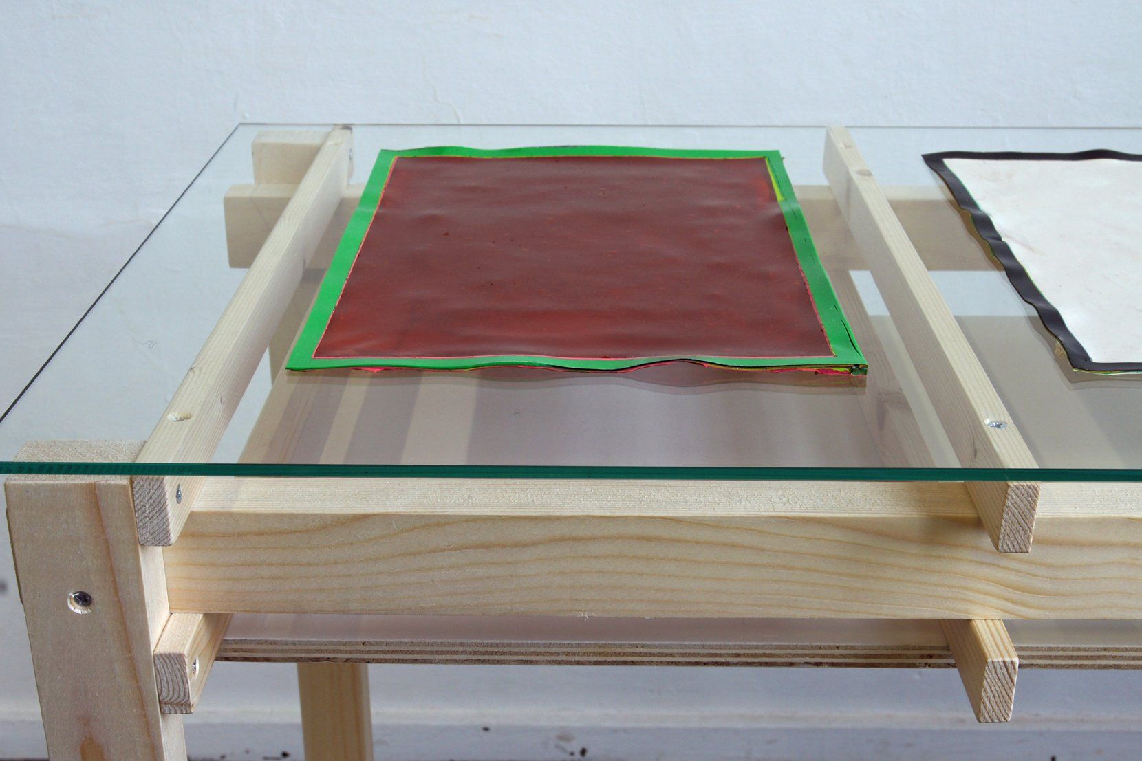 Op een houten tafel met een glazen blad ligt een rood rechthoekig voorwerp met een groene rand, naast een wit vel papier met een zwarte lijst.