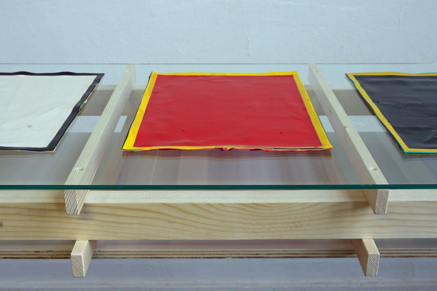 Een glazen tafelblad met drie rechthoekige panelen in wit, rood en zwart, rustend op een houten frame tegen een witte muur.