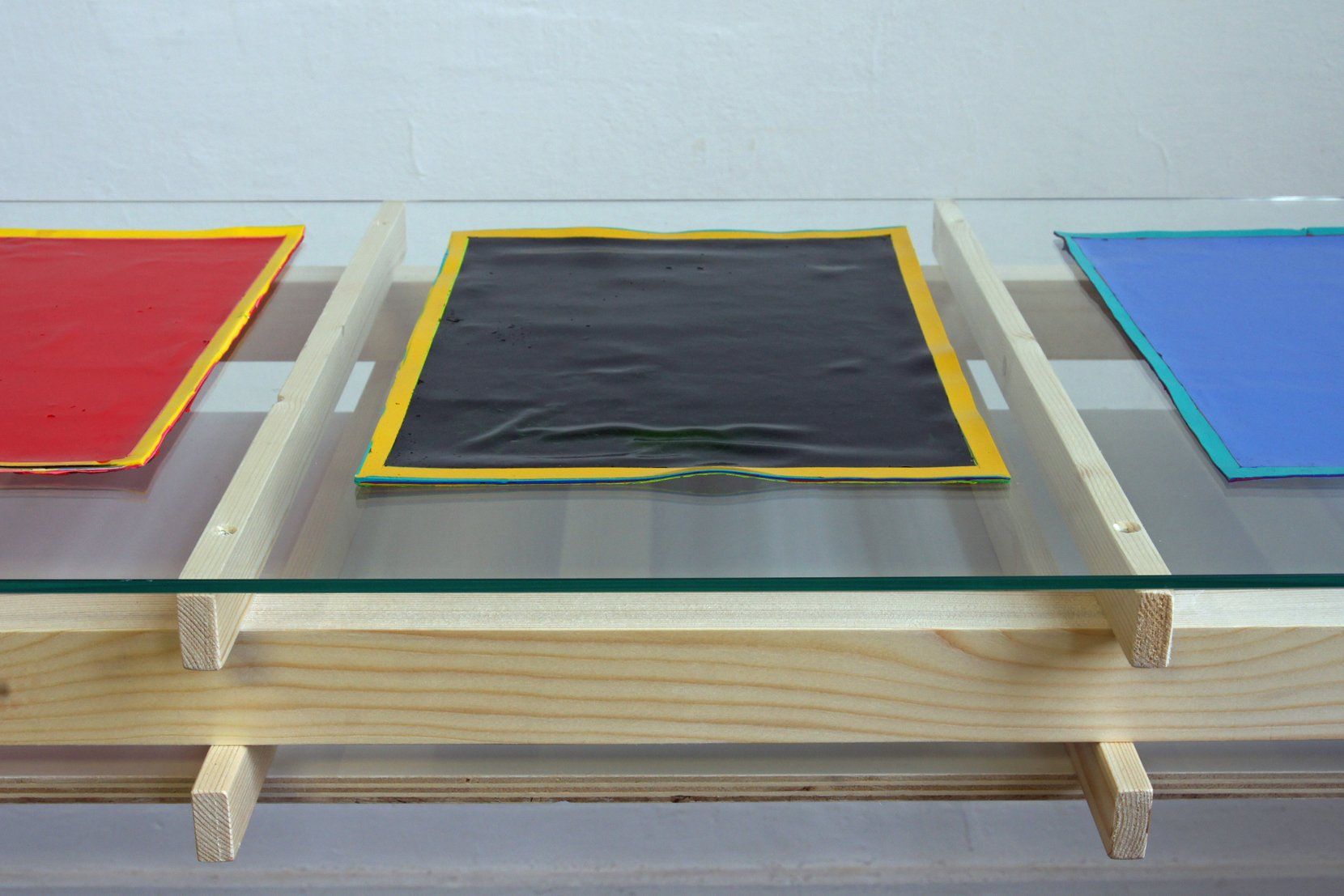 Op een glazen oppervlak, ondersteund door een lichtgekleurd houten frame, zijn een rood, een zwart en een blauw vierkant geplaatst.