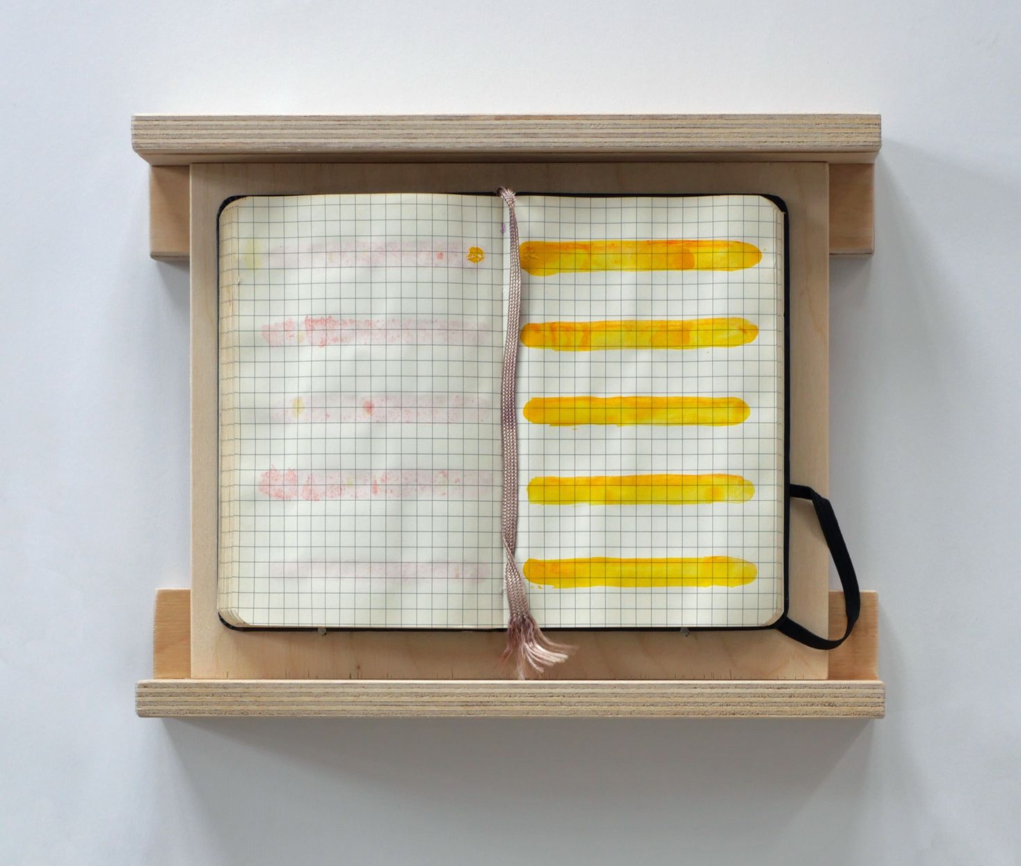 Een open notitieboekje ligt op een houten plank. De linkerpagina heeft vage roze vlekjes; de rechterpagina heeft dikke horizontale gele verfstrepen.