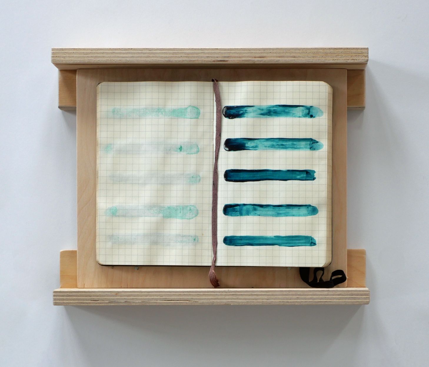 Open notitieboek met turquoise verfstreken, ingelijst in een houten frame aan een witte muur.