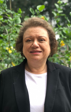 A woman in a black jacket and white shirt is standing in front of a tree.