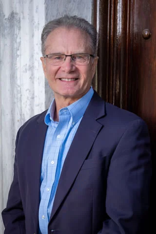 A man in a blue shirt and black pants is standing with his hands in his pockets.