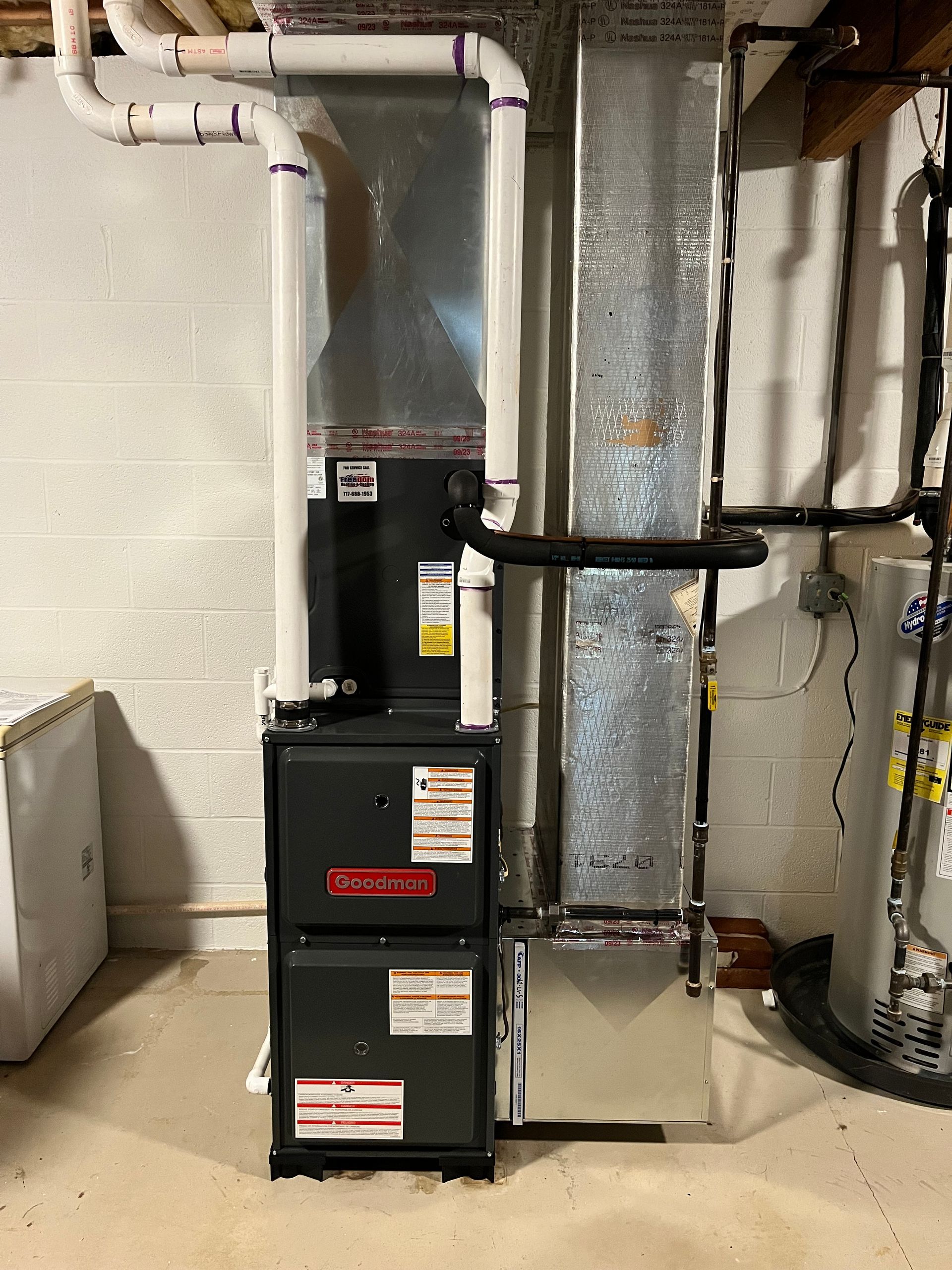 Furnace in a basement; black Goodman unit with silver ductwork and white PVC pipes.