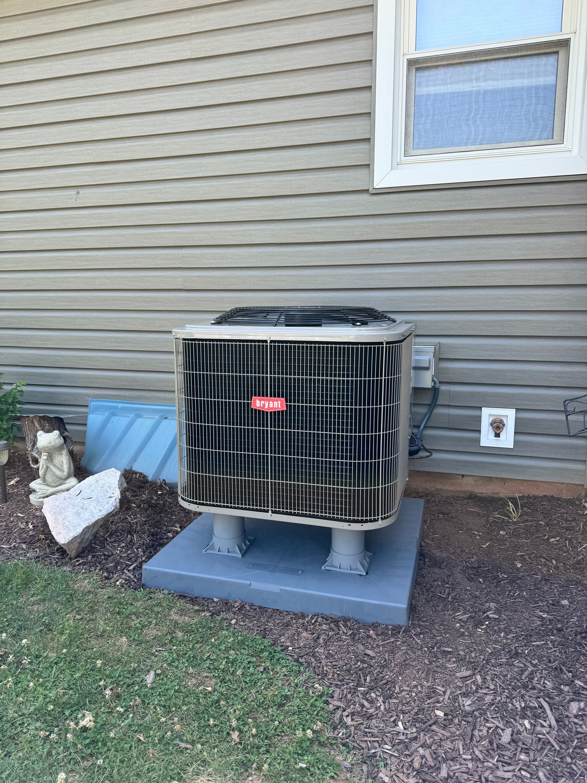 An air conditioning unit on a concrete pad near a building with siding, next to a window and landscaping.