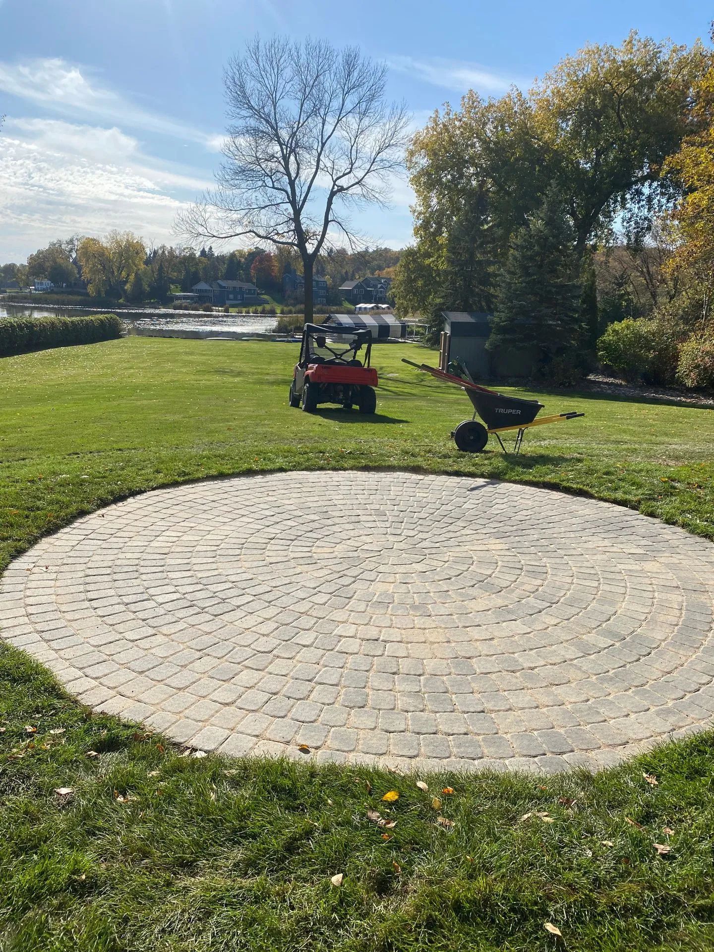 Circular stone patio in a sunny grassy park, with a red lawn mower and black wheelbarrow nearby.