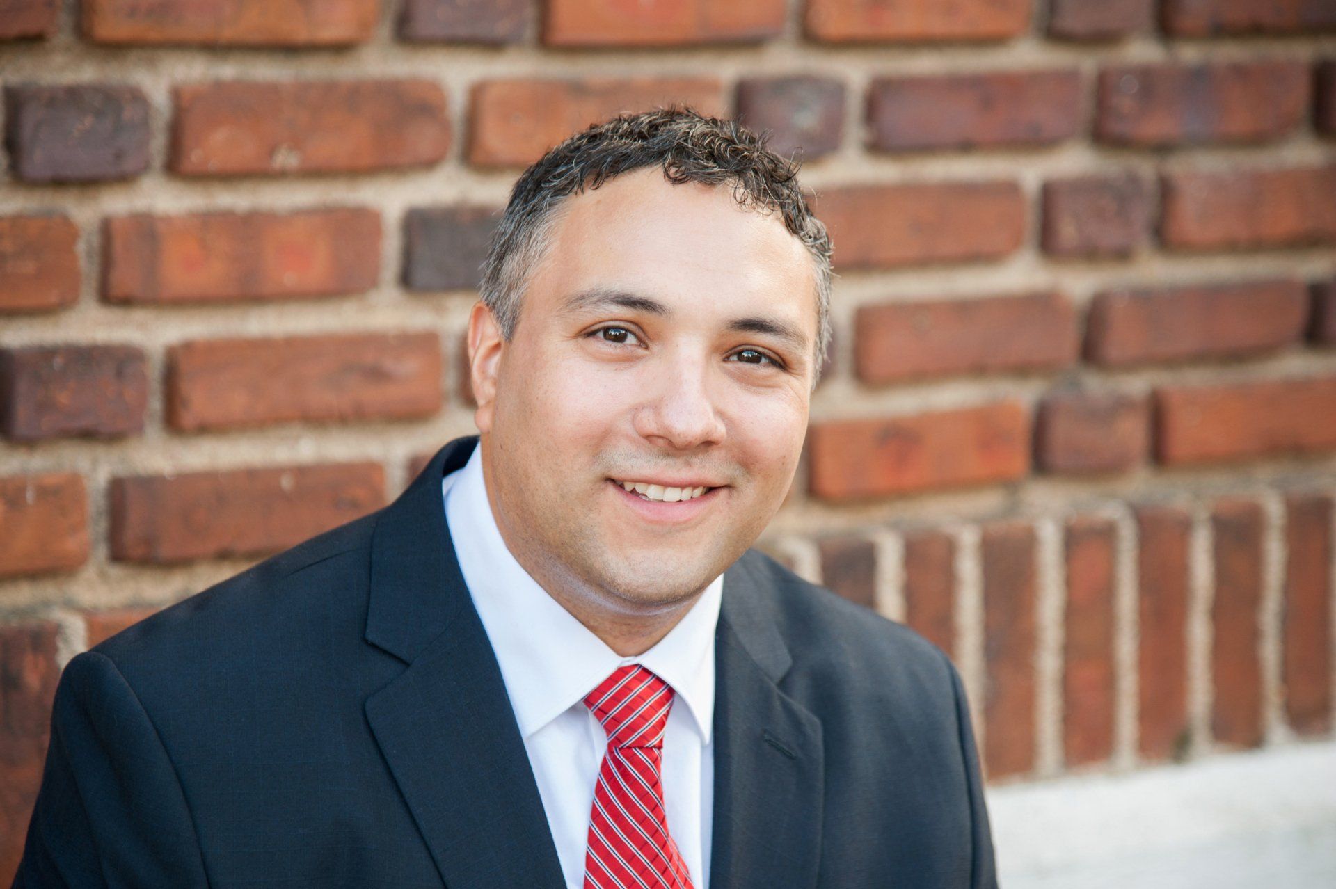 A man in a suit and tie is smiling in front of a brick wall.