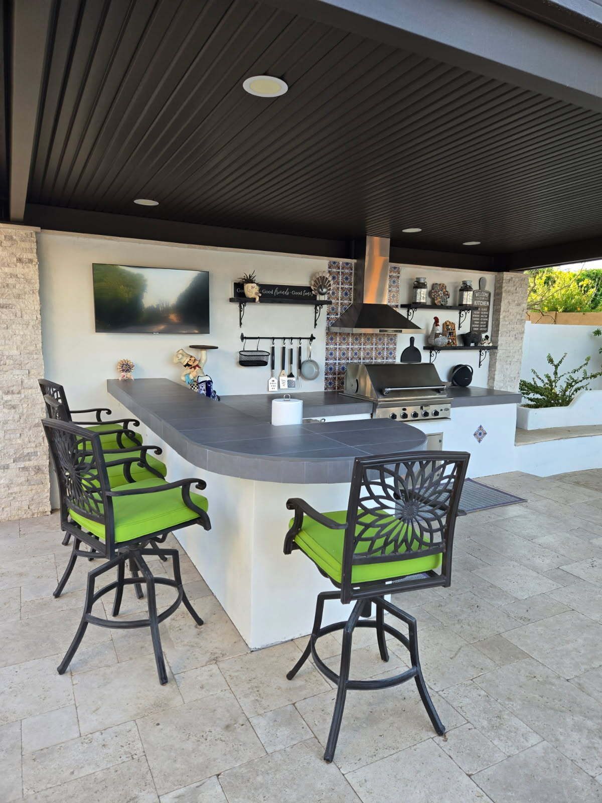 Outdoor covered bar with white counter, gray top, green stools, and shelves with glasses and bottles