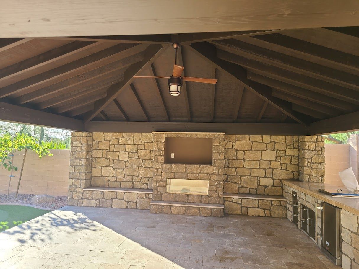 Stone outdoor pavilion with a dark wooden roof, built-in grill, and bench seating.