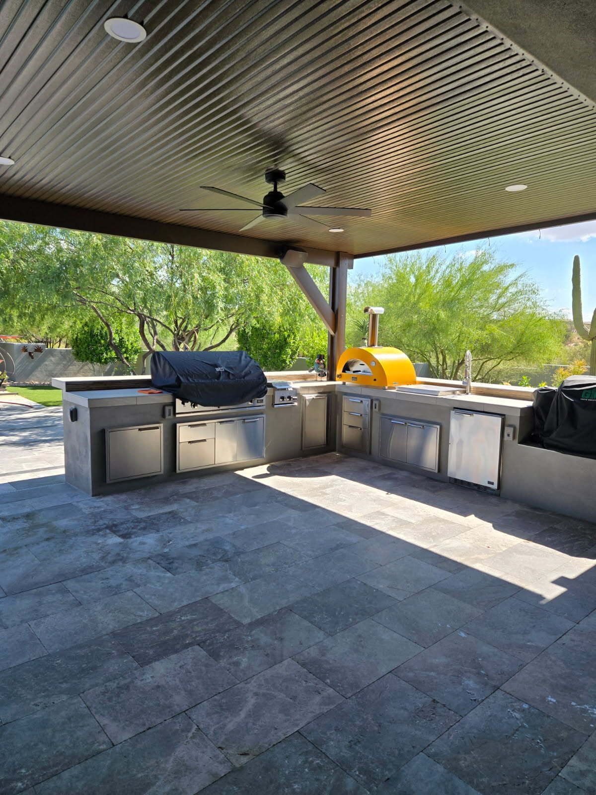 Open-air patio with built-in grill, counter, and sinks under a shaded roof, overlooking trees and desert hills