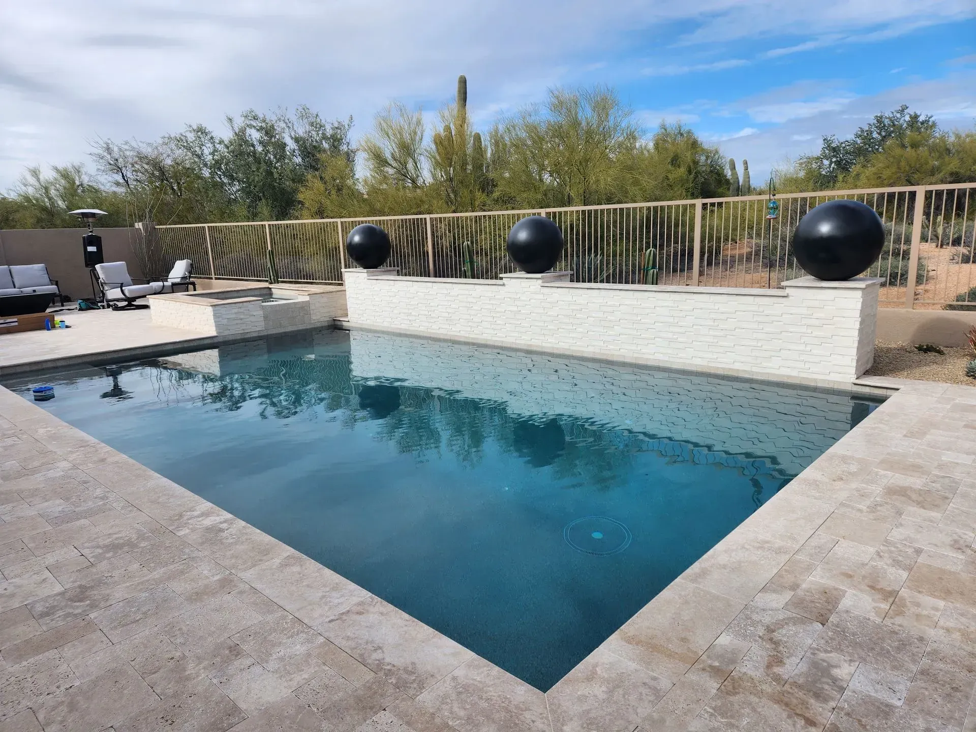 Modern backyard pool with stone deck, lounge chairs, and black spherical planters by a white wall
