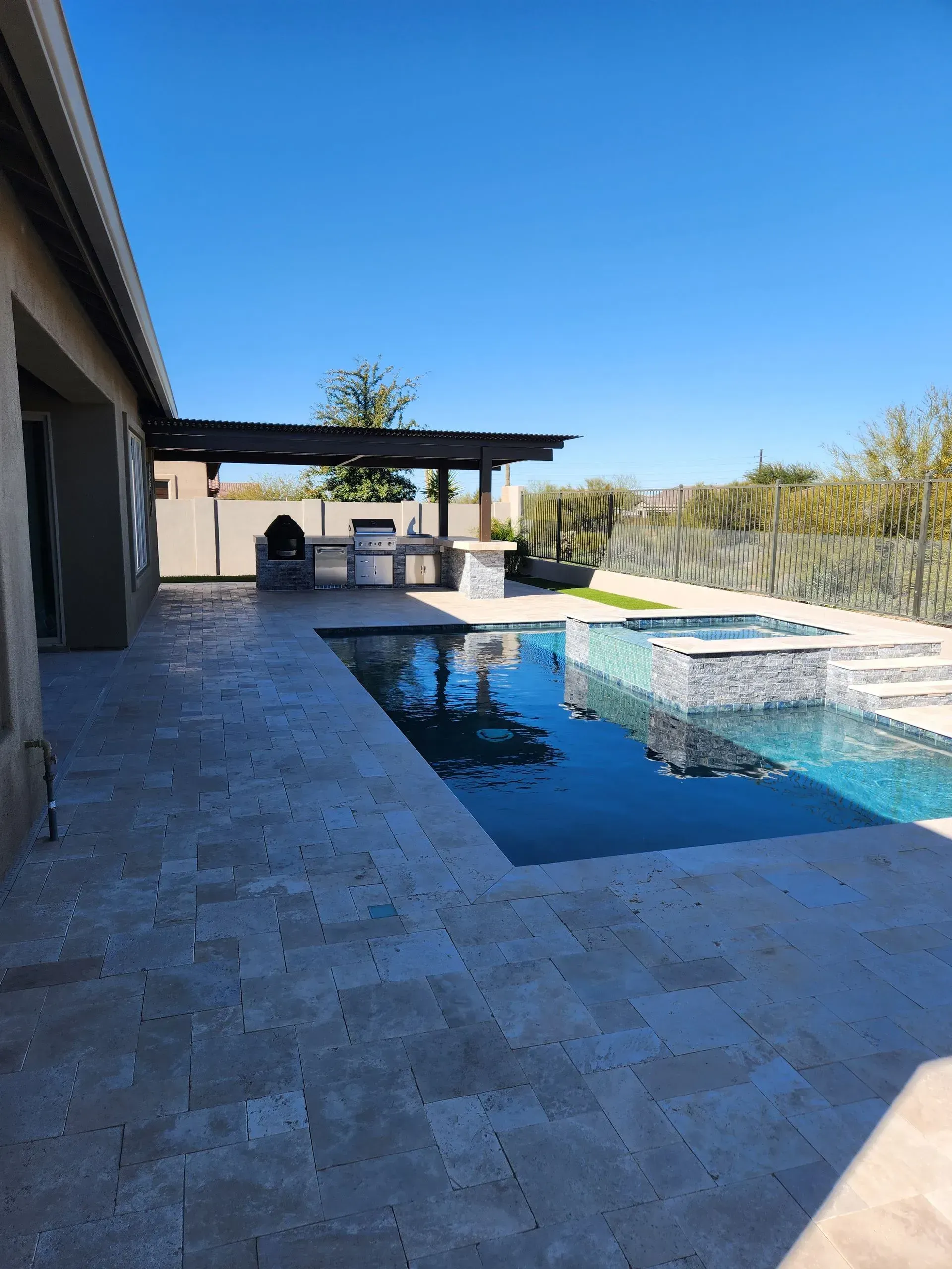 Stone patio with a modern backyard pool, covered seating area, and bright blue sky.