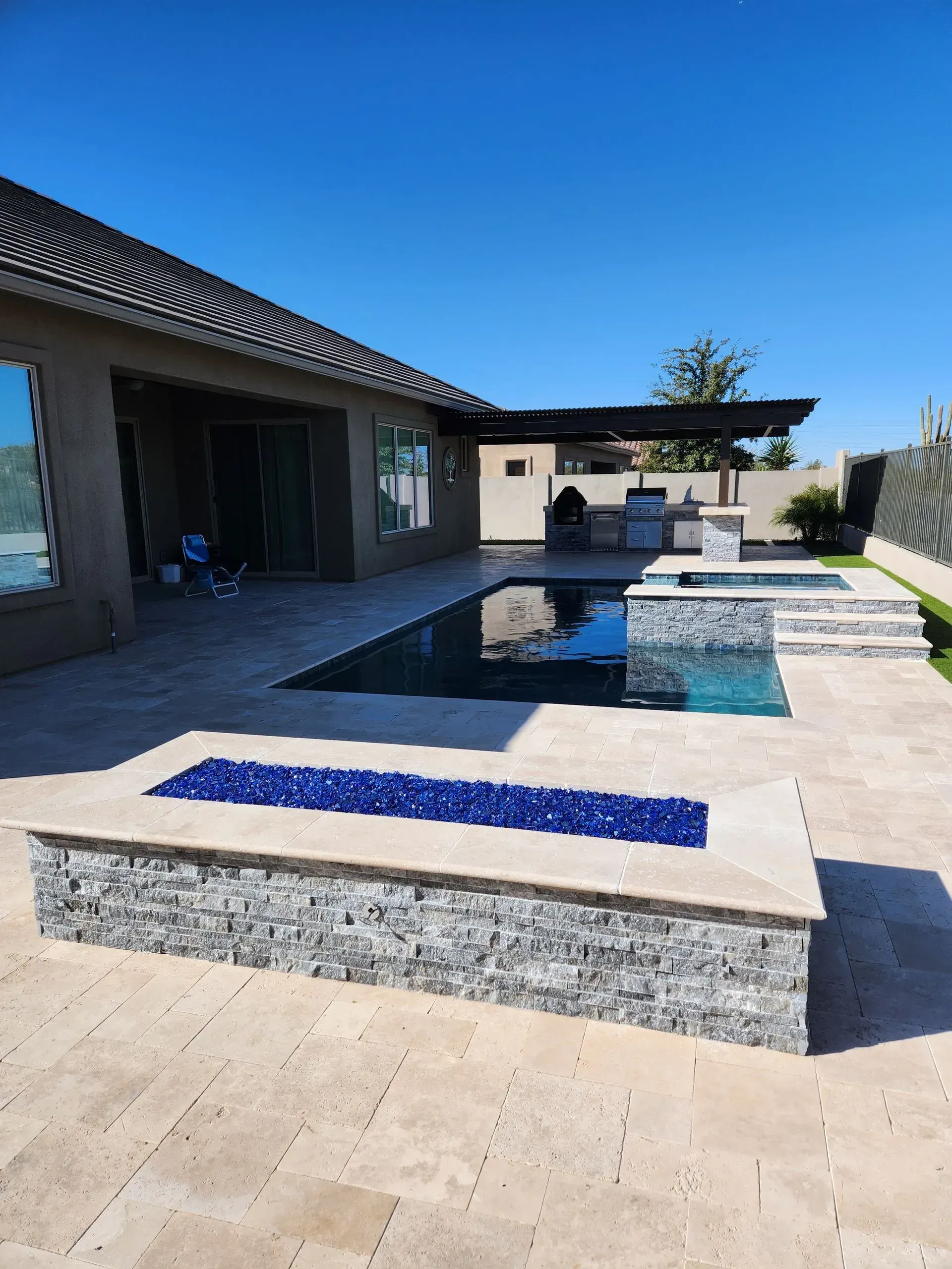 Modern backyard with a blue-tiled pool, beige lounge beds, and a shaded patio beside a house