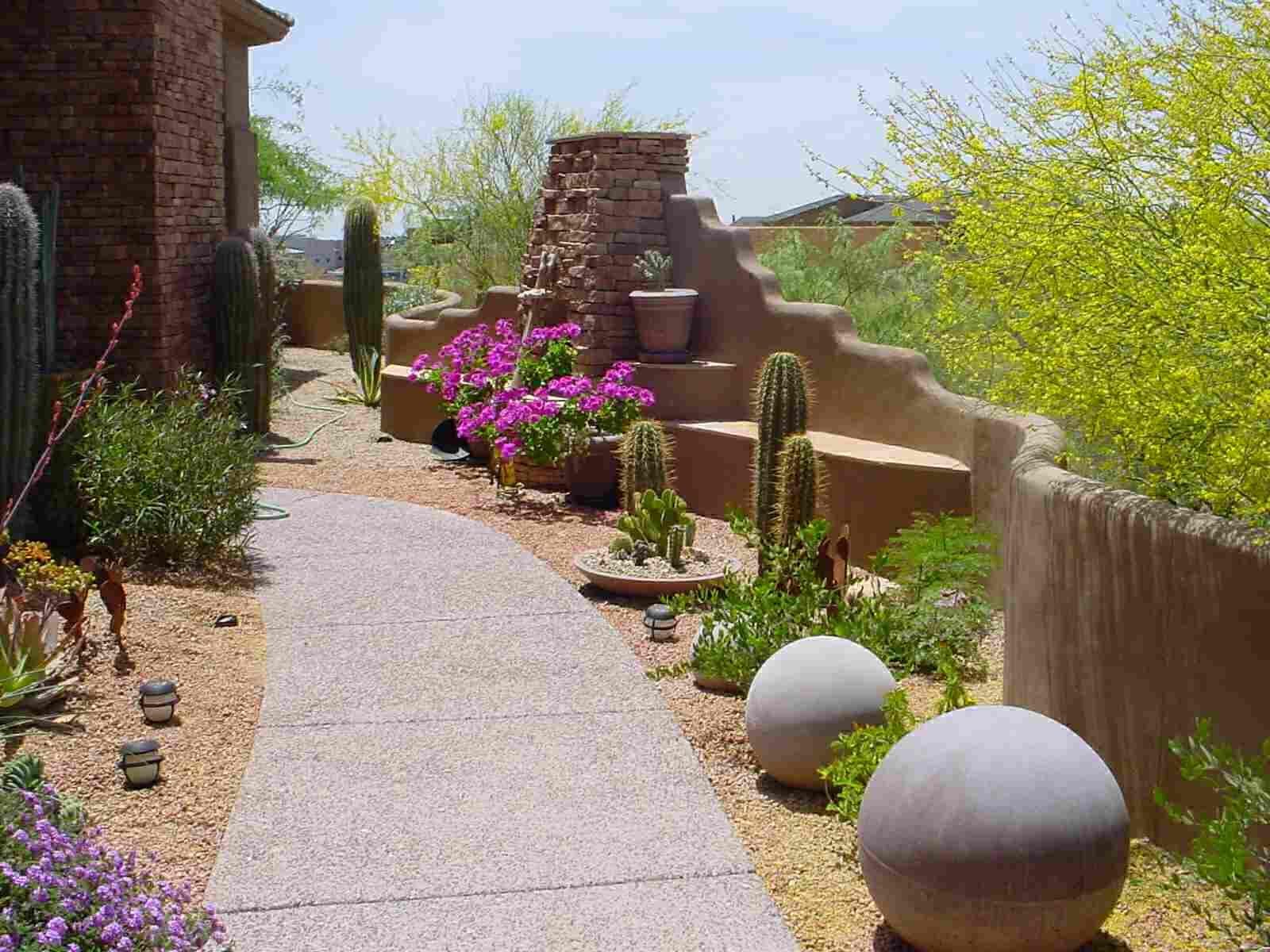 Curved desert garden path with pink flowers, cacti, and large decorative stone spheres beside a stucco wall