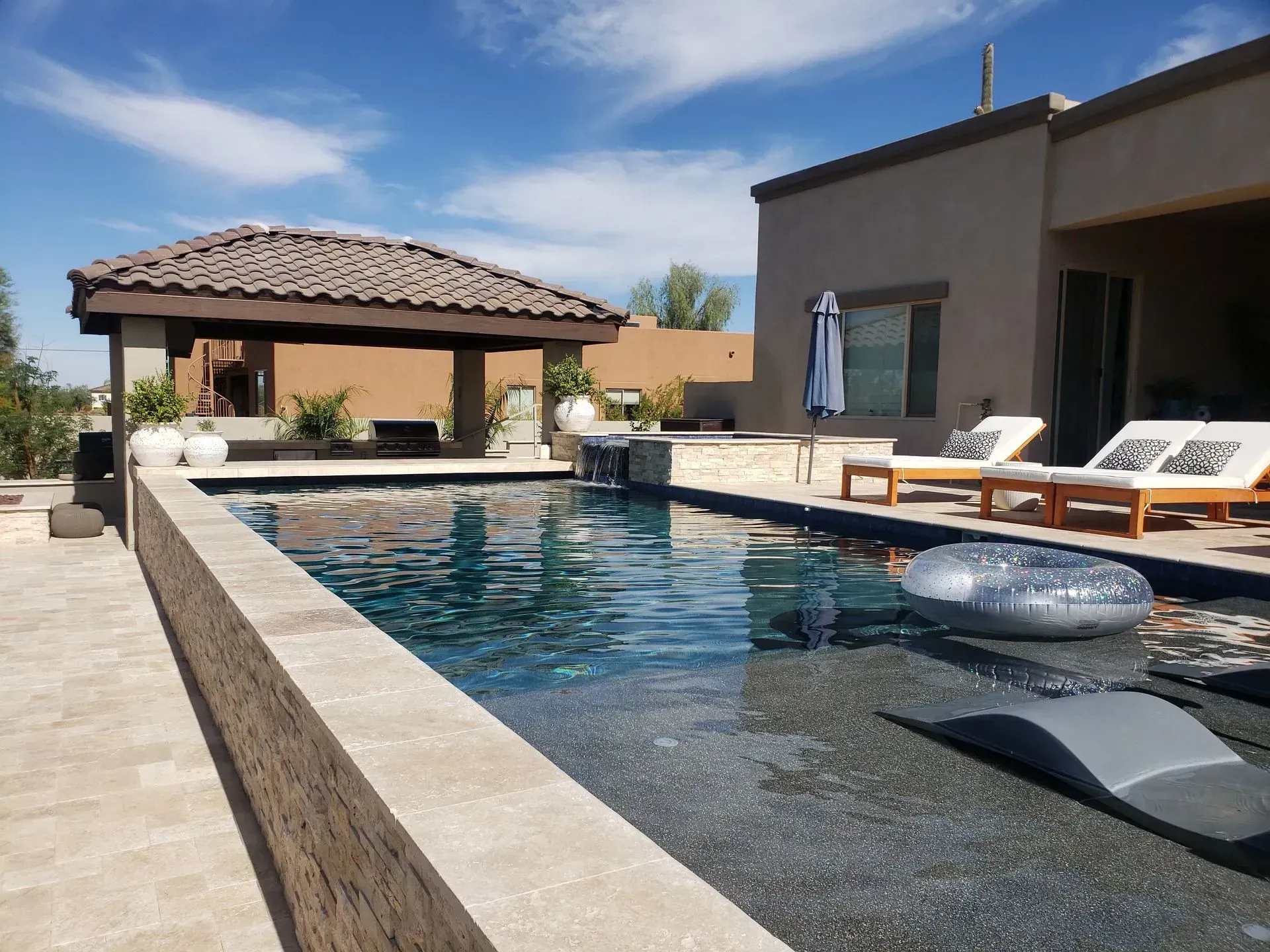 Modern backyard pool with lounge chairs, shaded gazebo, and a desert-style house under a clear blue sky