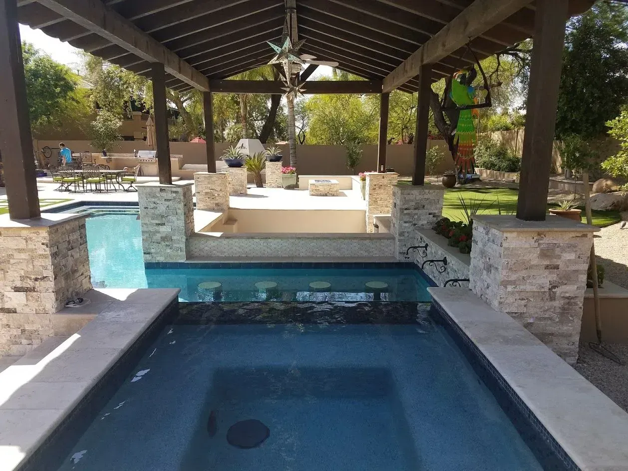Covered patio with a small rectangular pool and stone bar seating area, surrounded by greenery.