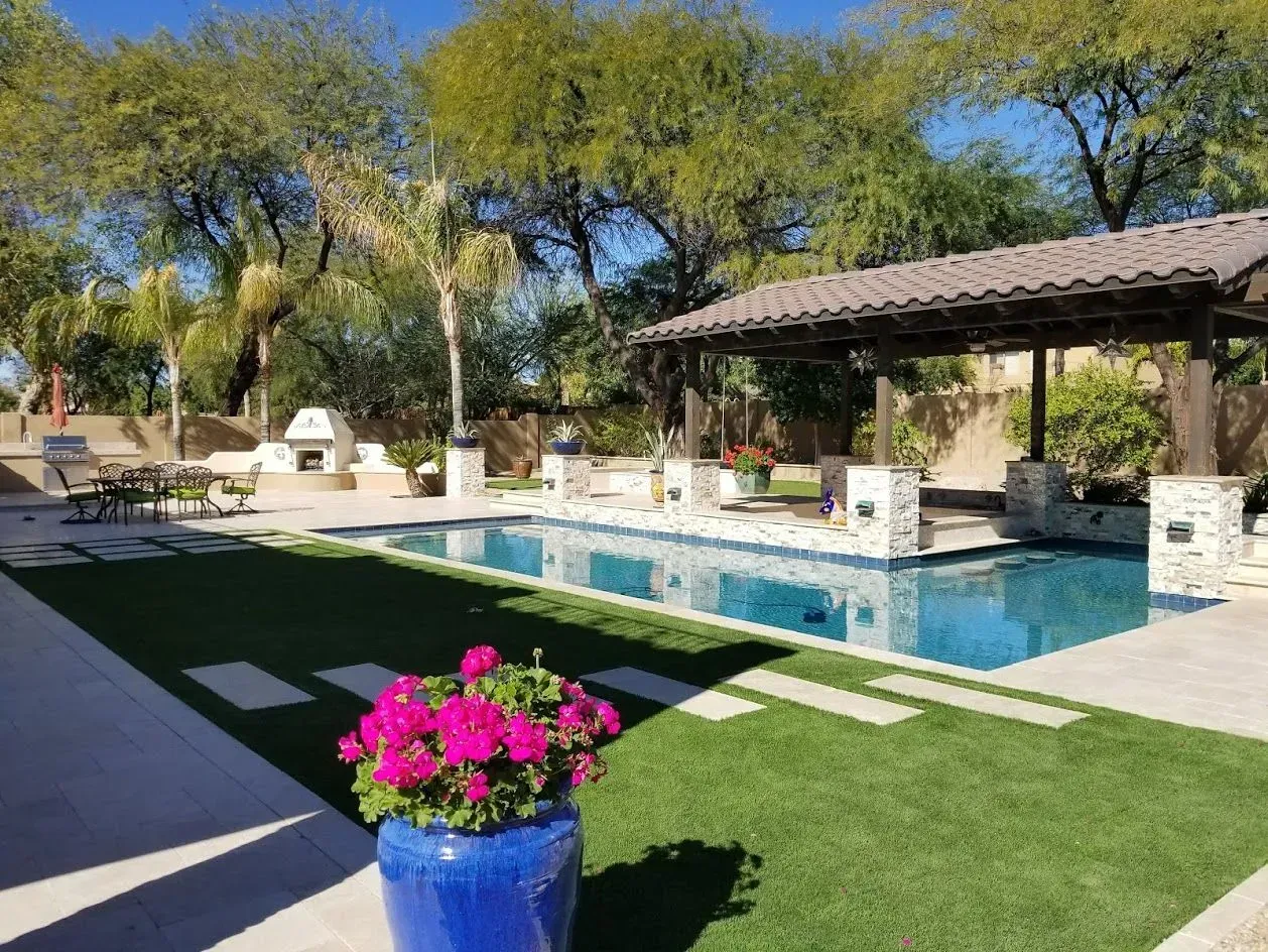 Backyard pool with stone patio, shaded gazebo, and bright pink flowers in a blue pot