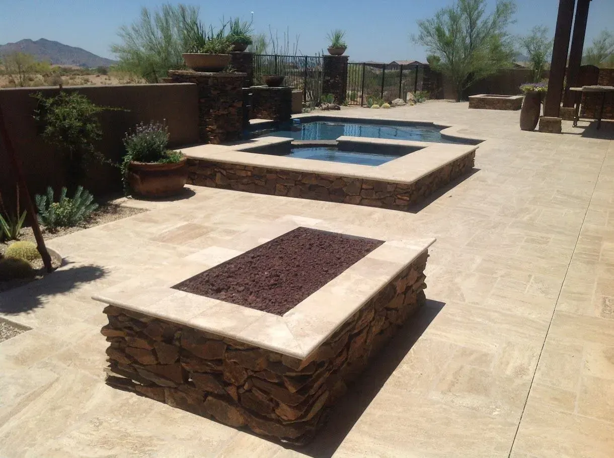 Desert patio with stone fire pit, raised spa, and tan paver deck under a clear sky
