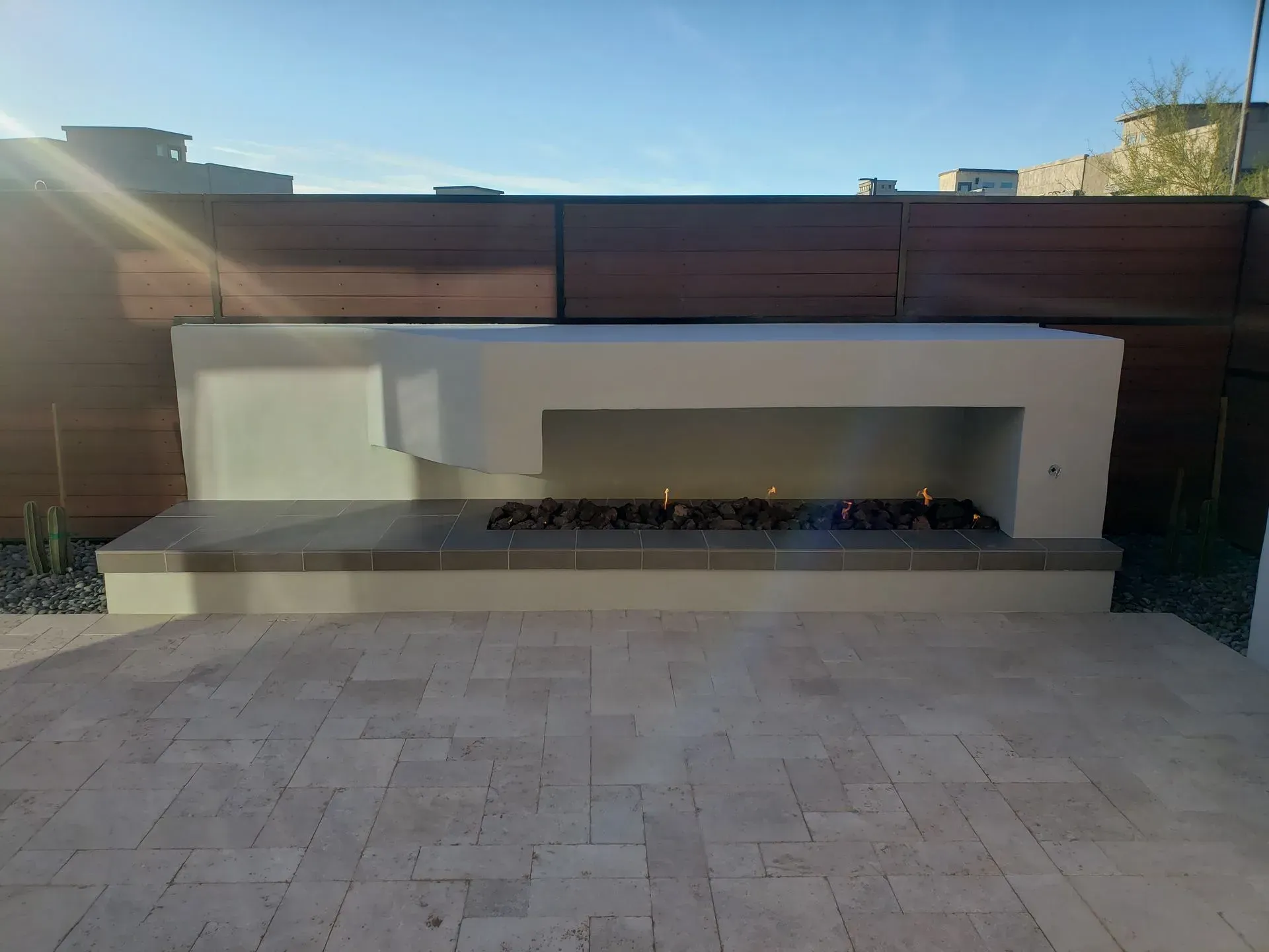 Modern outdoor fireplace and seating wall on a paved patio, set against a wooden fence under blue sky