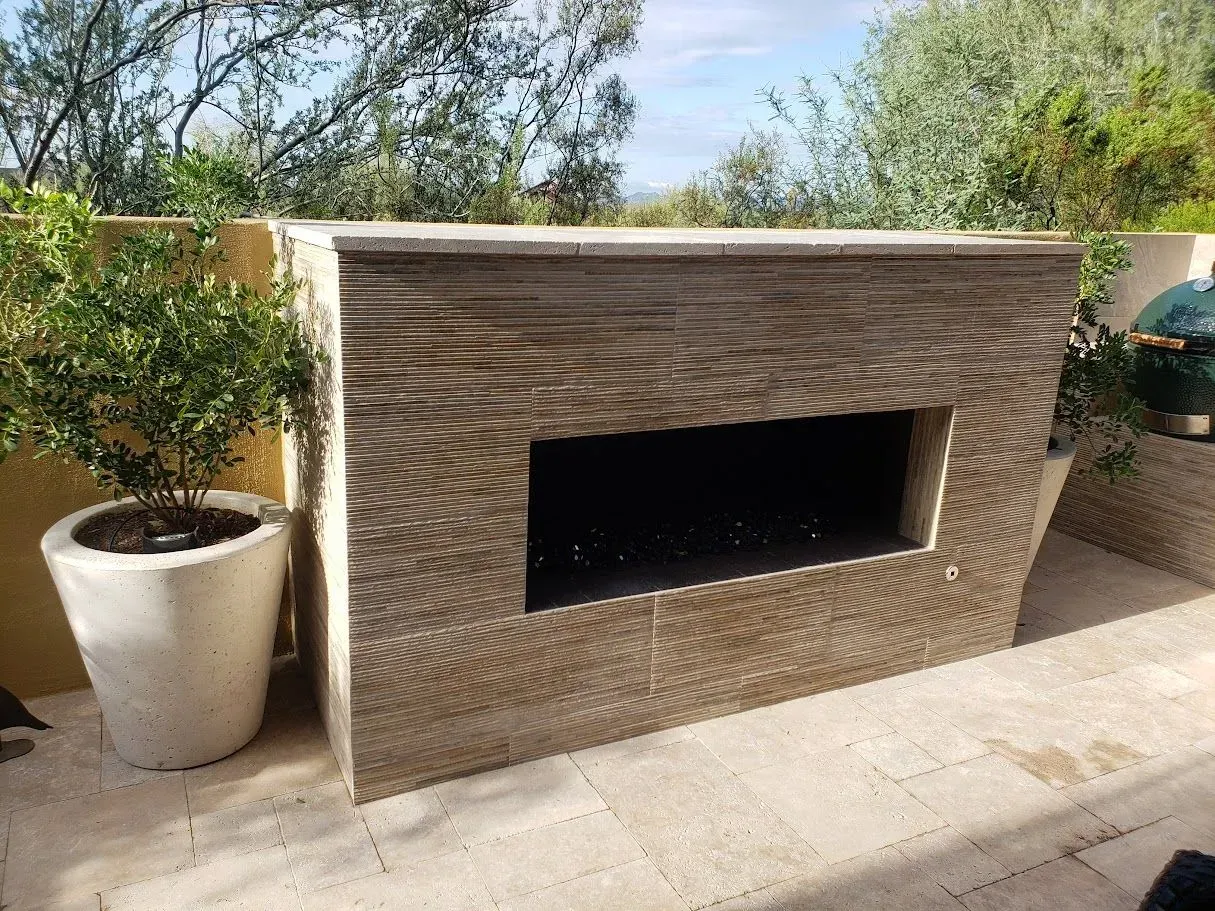 Outdoor brick fireplace with a wide rectangular opening, flanked by potted plants on a patio.