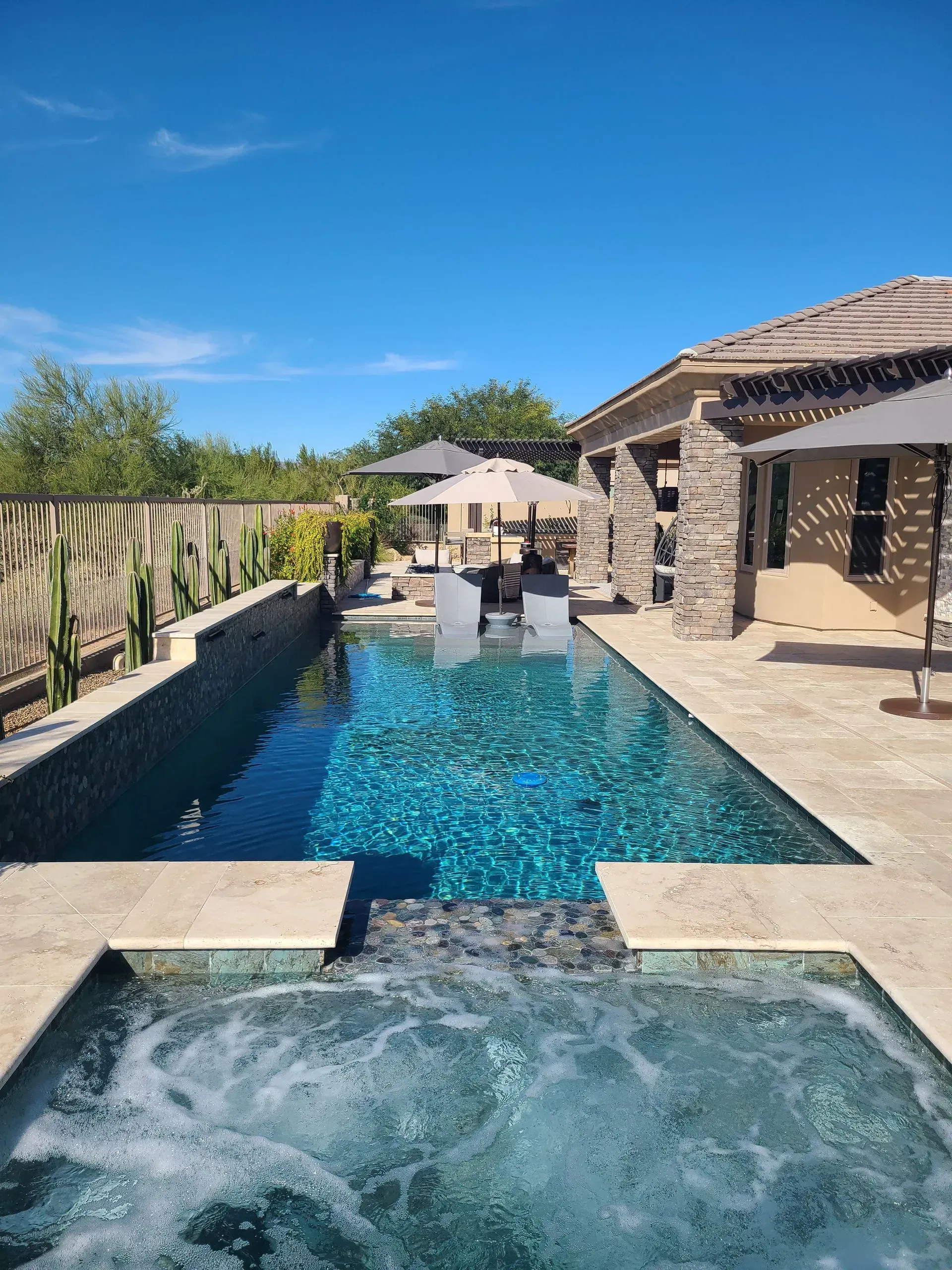 Backyard pool with hot tub, stone patio, and a stone house under a bright blue sky