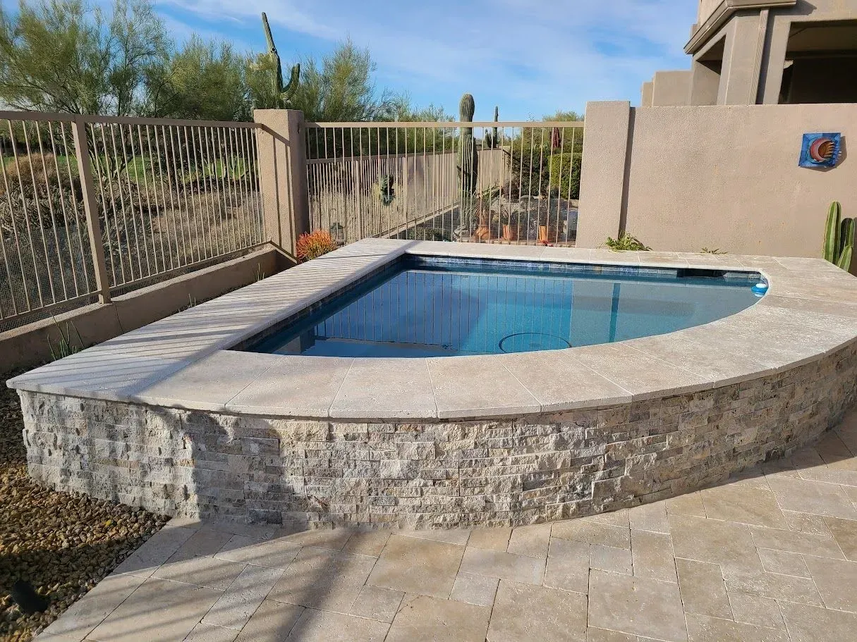 Small outdoor stone pool with blue water beside a paved patio and fence under a clear sky