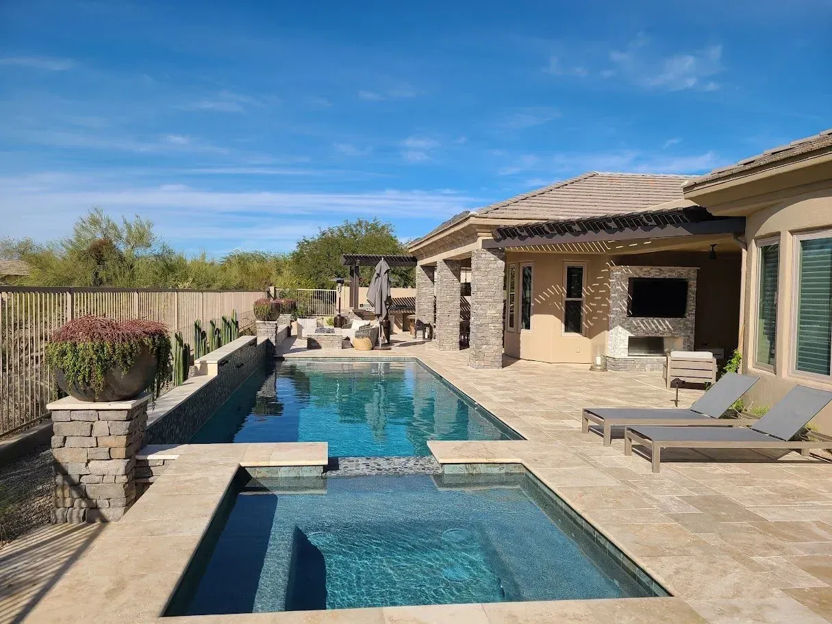 Backyard patio with a blue pool, spa, lounge chairs, and a stone house under a clear sky