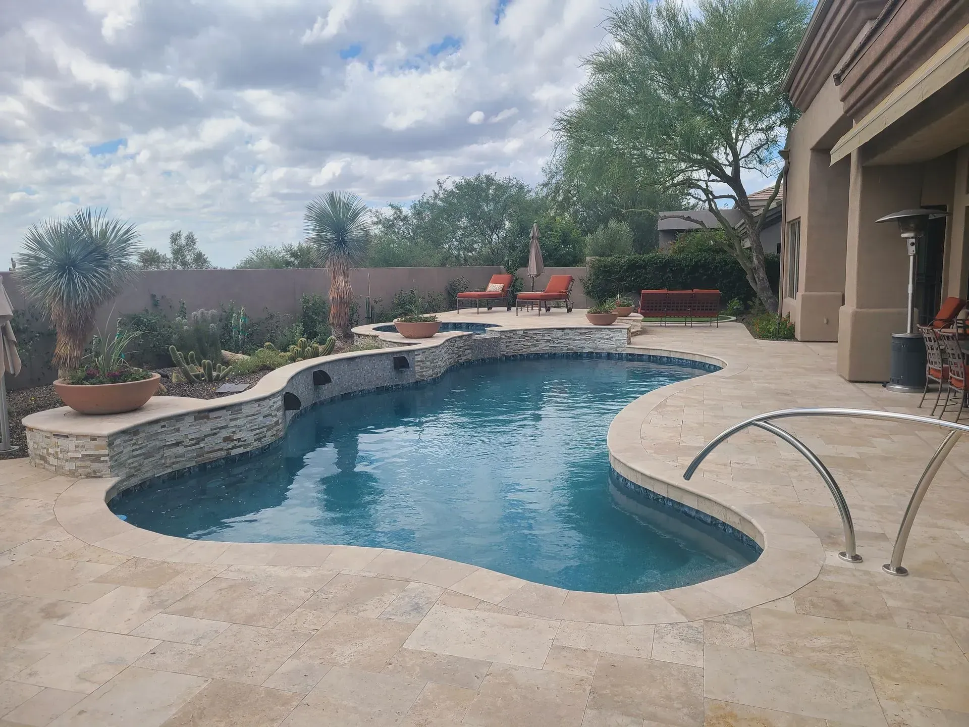 Backyard swimming pool with stone deck, patio seating, and desert landscaping under a cloudy sky