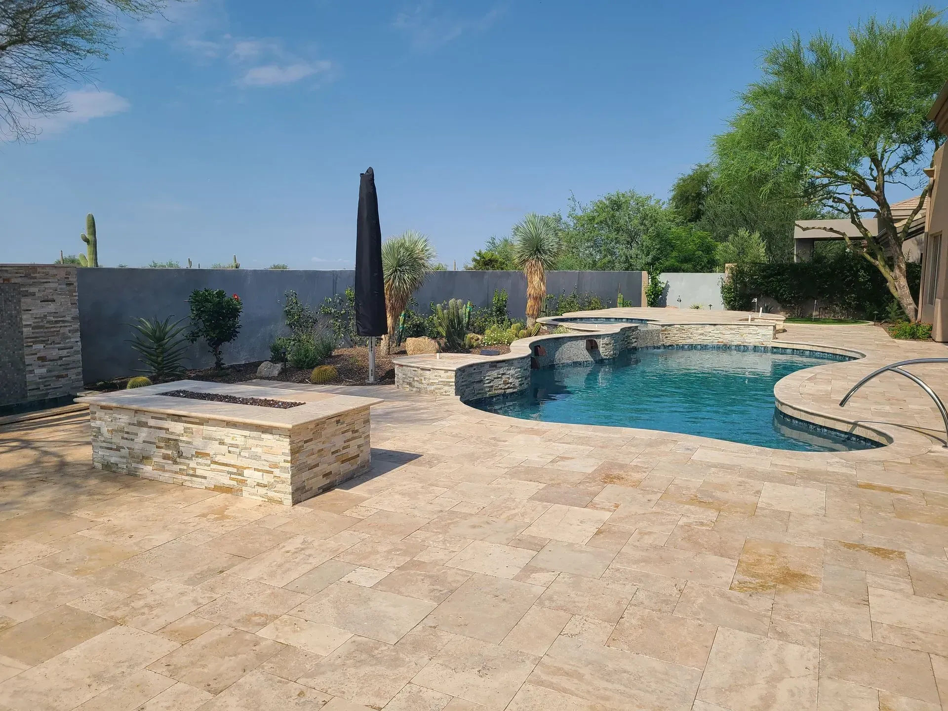 Stone patio with curved swimming pool, lounge chairs, and greenery under a clear blue sky