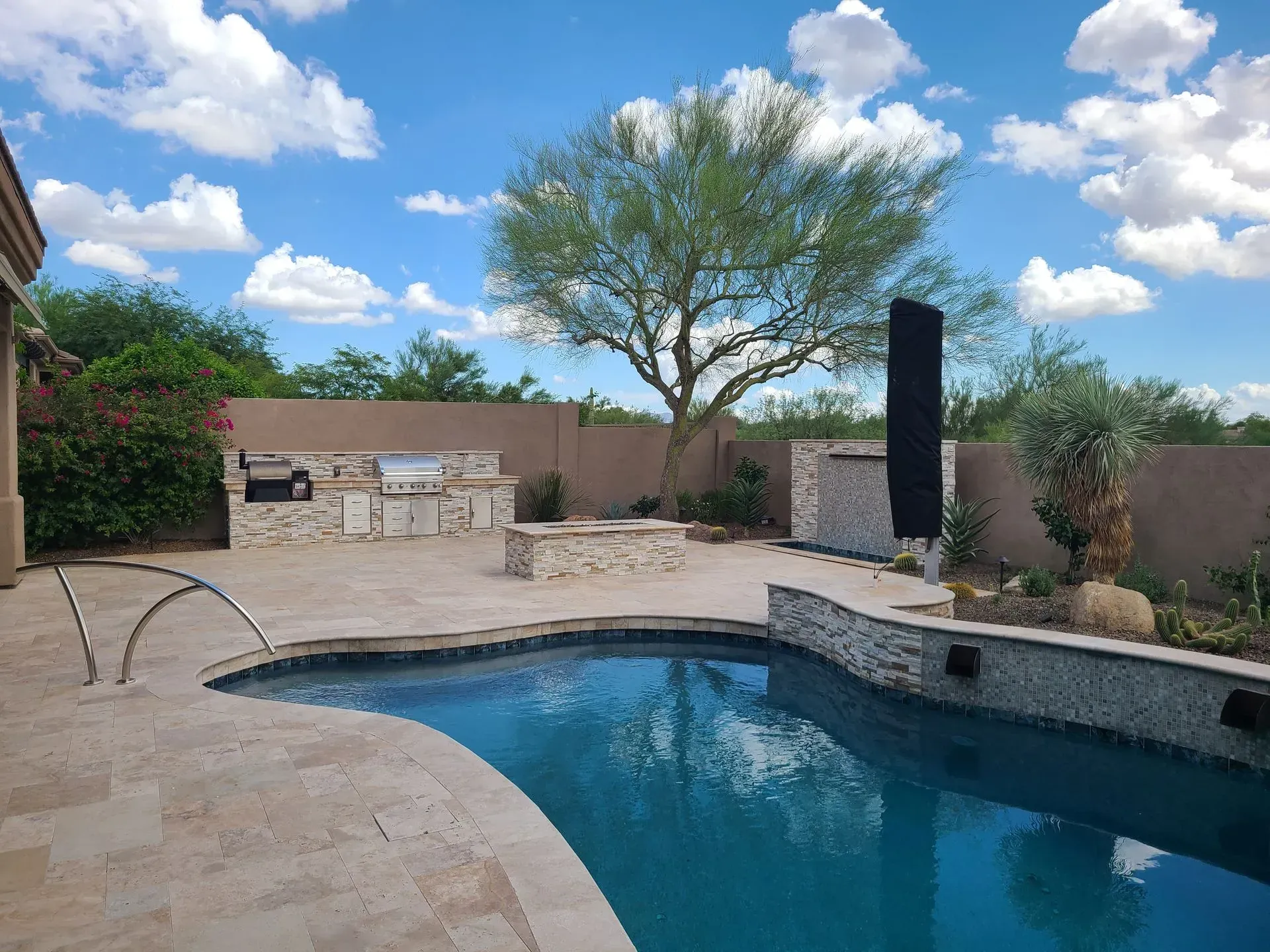 Backyard pool with blue water, stone patio, outdoor seating, and desert landscaping under a partly cloudy sky