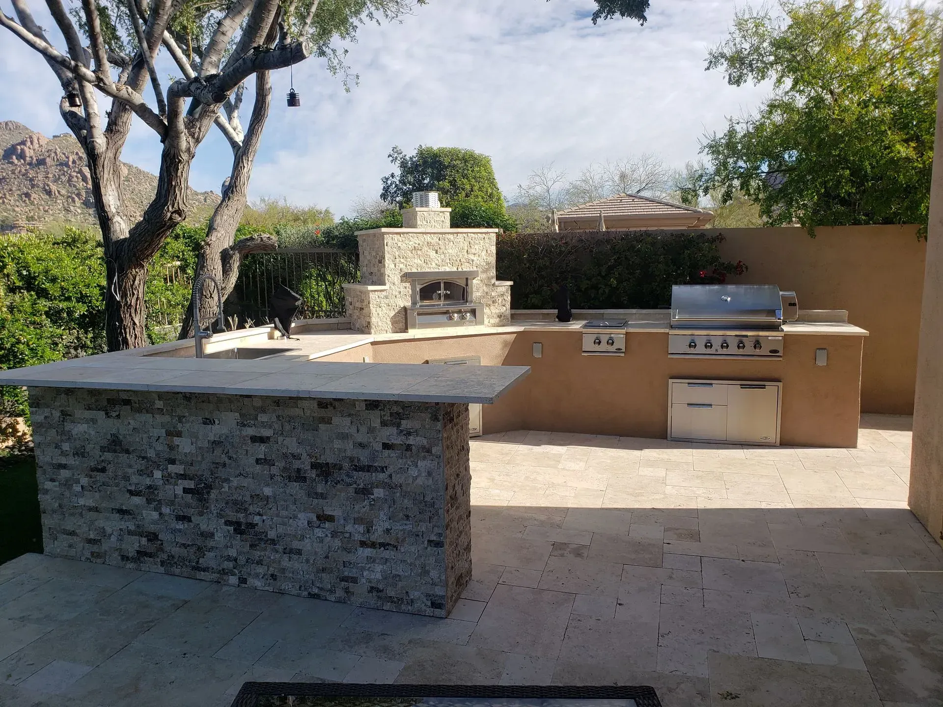 Backyard outdoor kitchen with stone island, built-in grill, and brick pizza oven in sunlight