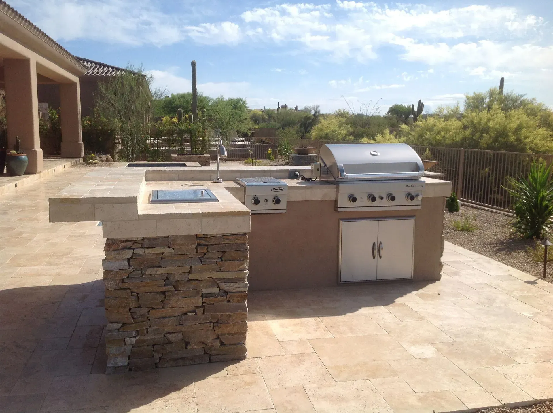 Outdoor stone patio with built-in grill, side counter, and landscaped yard under a bright sky