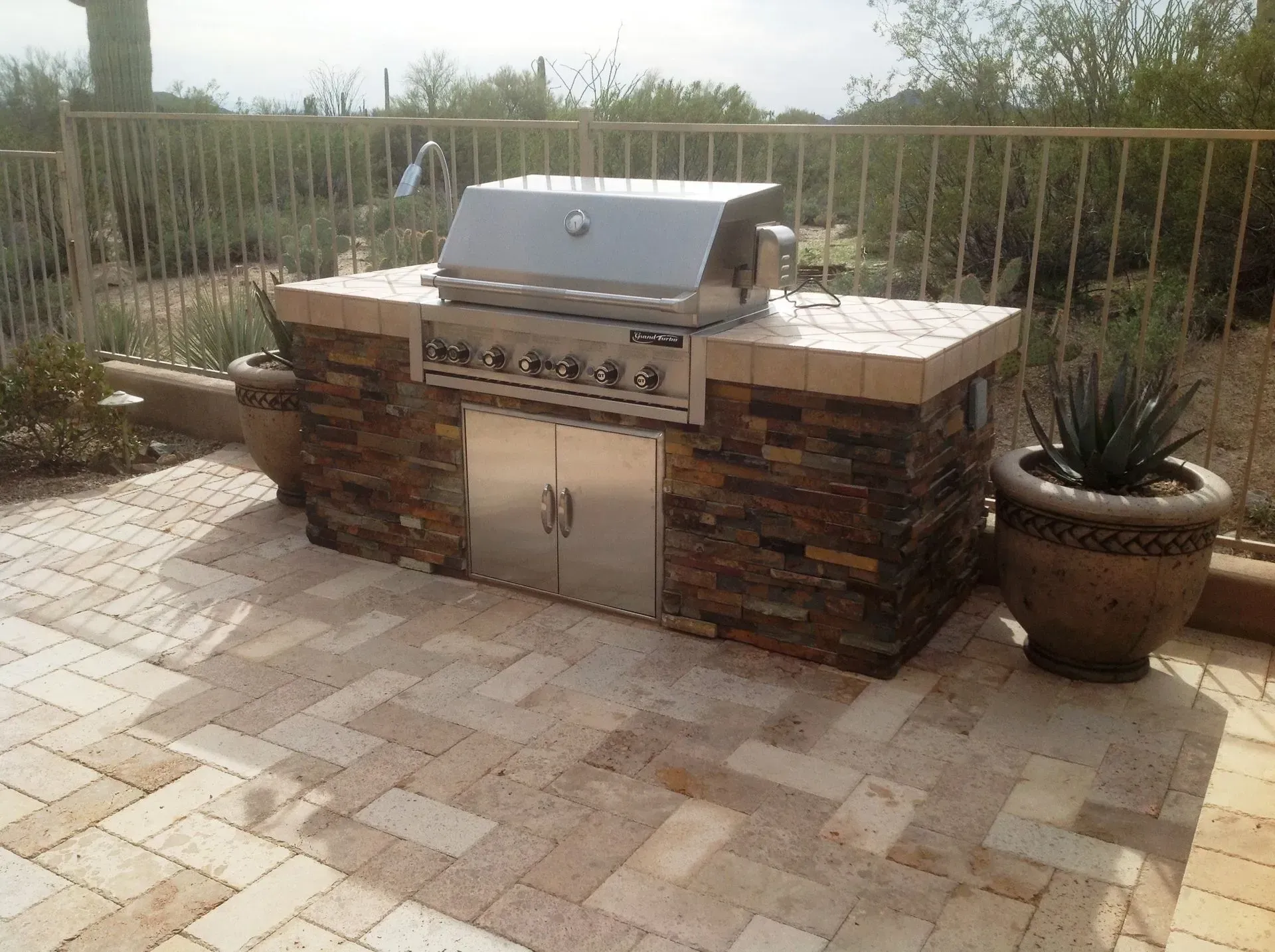 Outdoor stainless steel grill with stone base on a brick patio, flanked by two potted plants