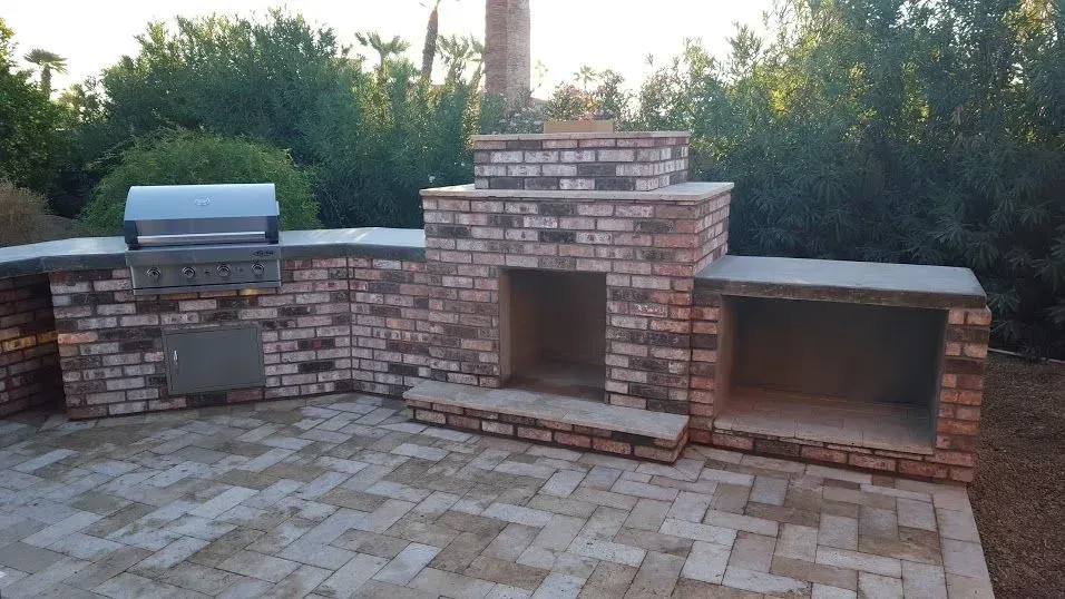 Brick outdoor fireplace and built-in grill on a paved patio, with trees in the background