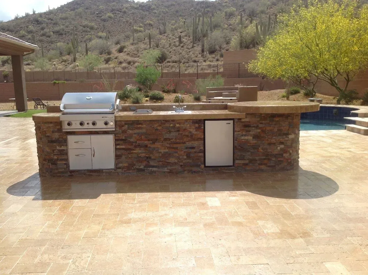 Outdoor stone barbecue island beside a pool, with desert hills and a tree in the background