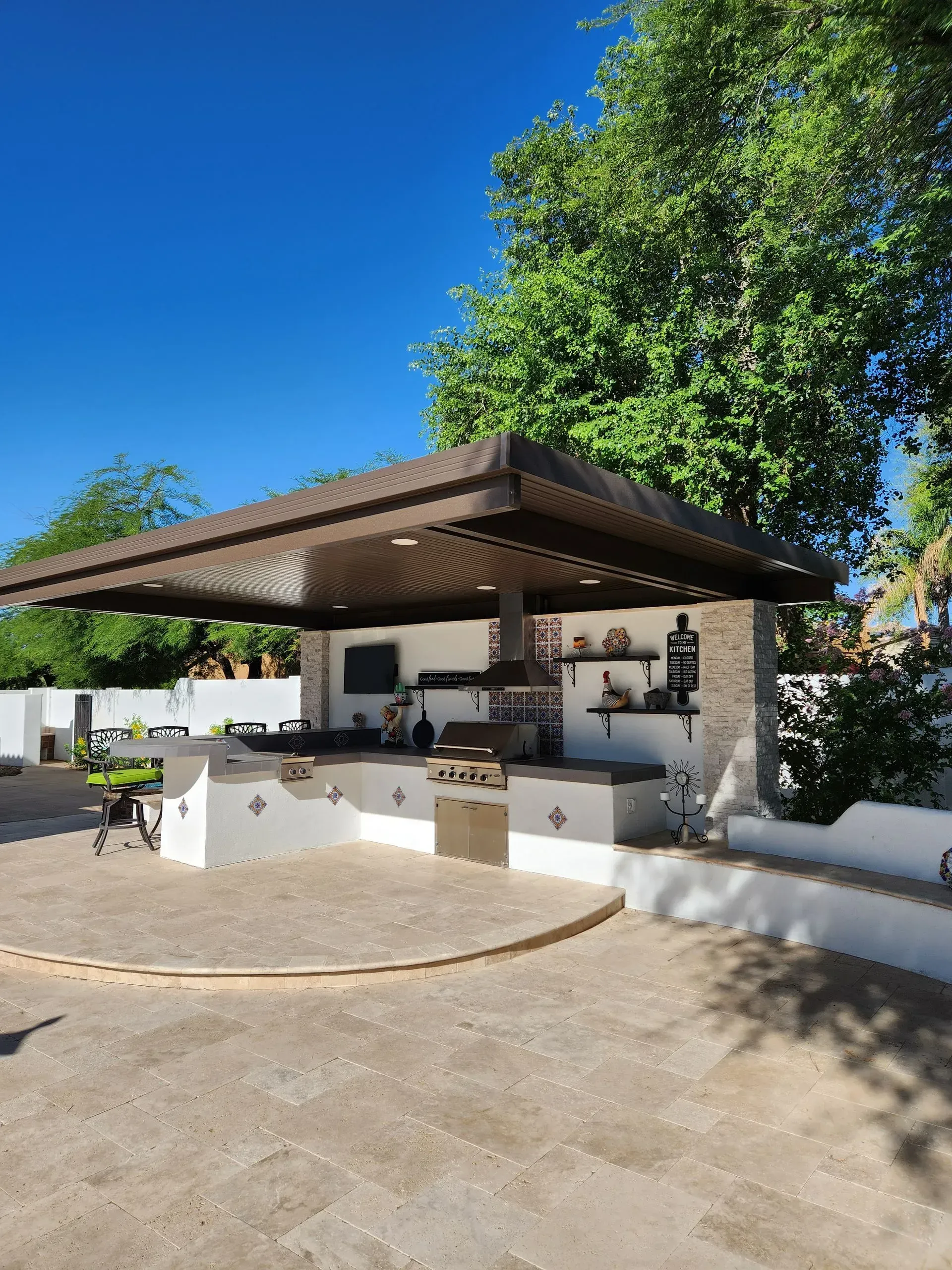 Outdoor stone bar with a wooden roof, white counters, and stools under a large tree in a sunny courtyard