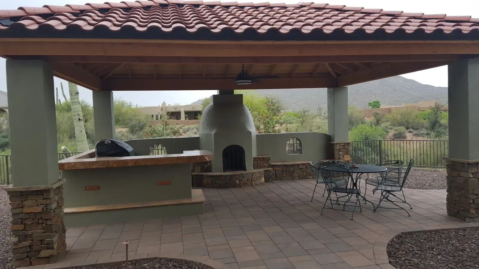 Outdoor patio with a tiled roof, stone fireplace, picnic table, and metal chairs overlooking a yard