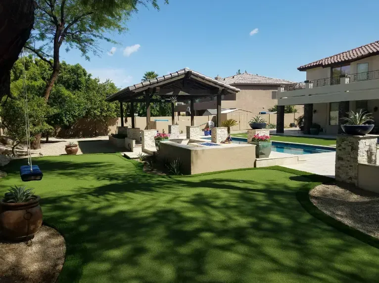 Backyard patio with artificial grass, a pergola, lounge chairs, and a swimming pool under a sunny sky
