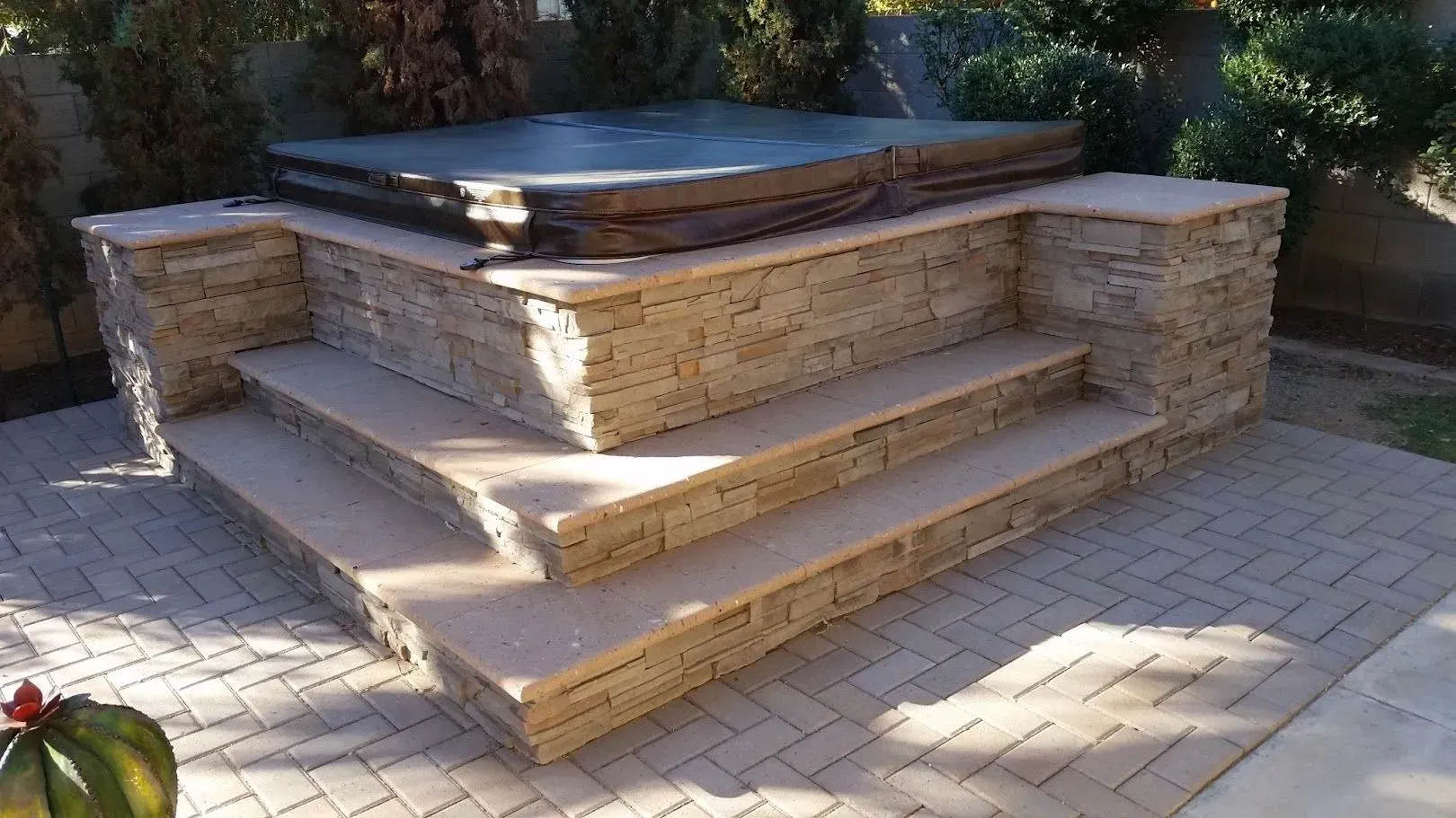 Stone steps and platform with seating around a raised wooden hot tub in a sunlit patio area