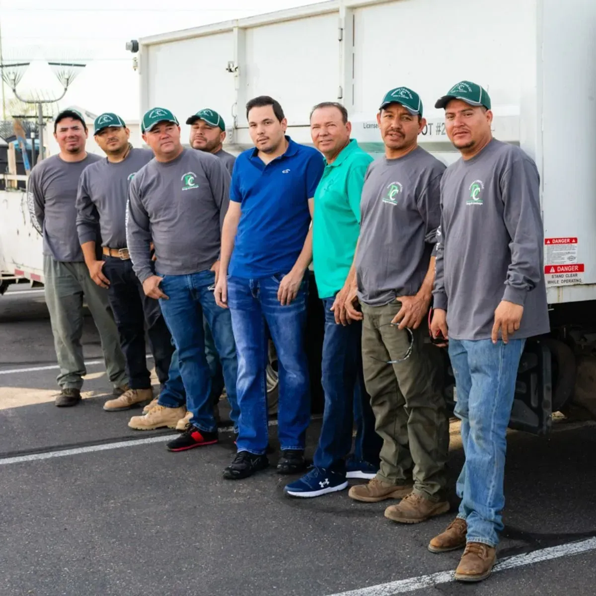 Seven workers standing in a line outdoors beside a white truck, wearing gray shirts and teal caps.