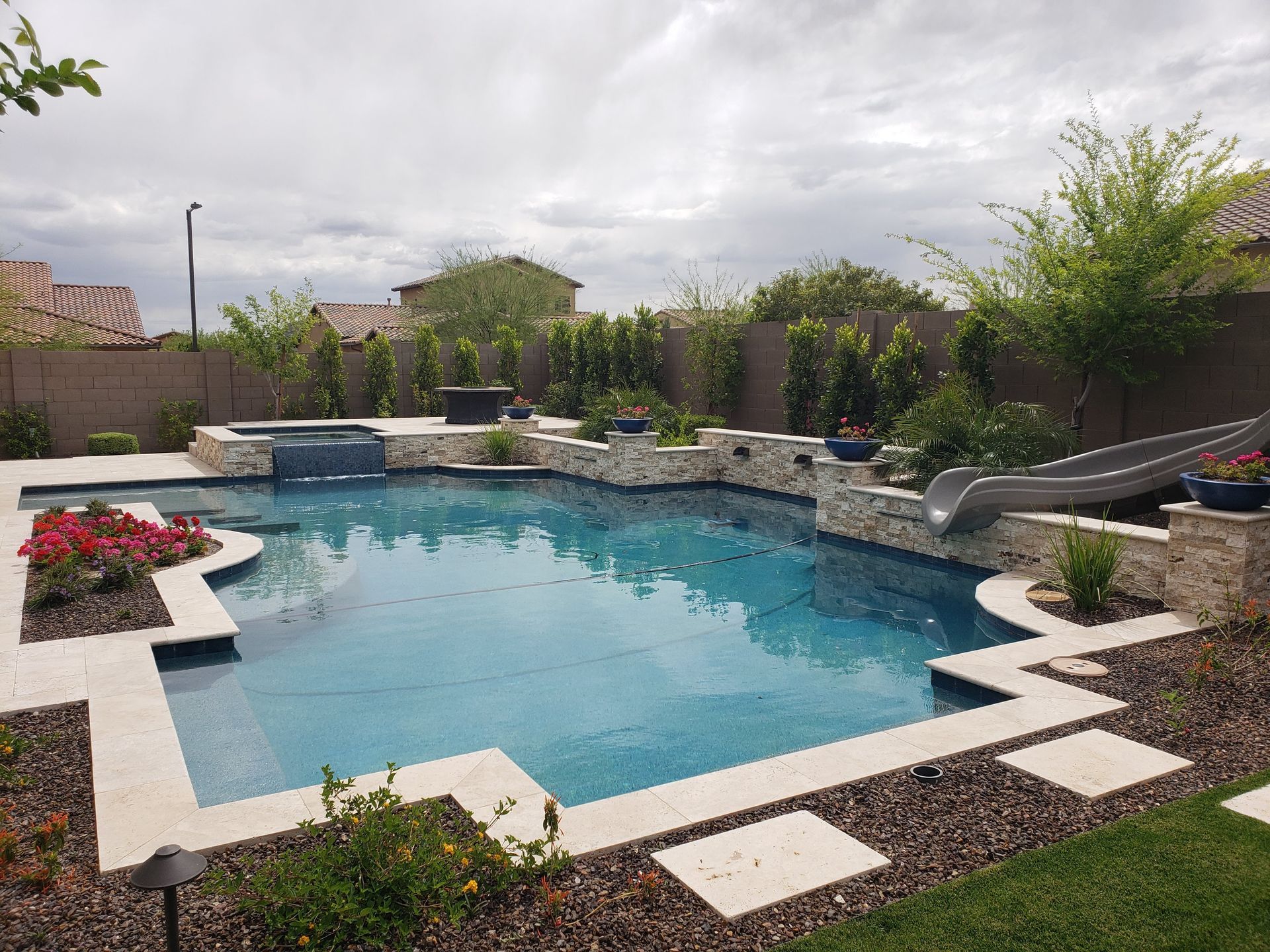 Swimming pool with stone features, slide, and landscaping under a cloudy sky.