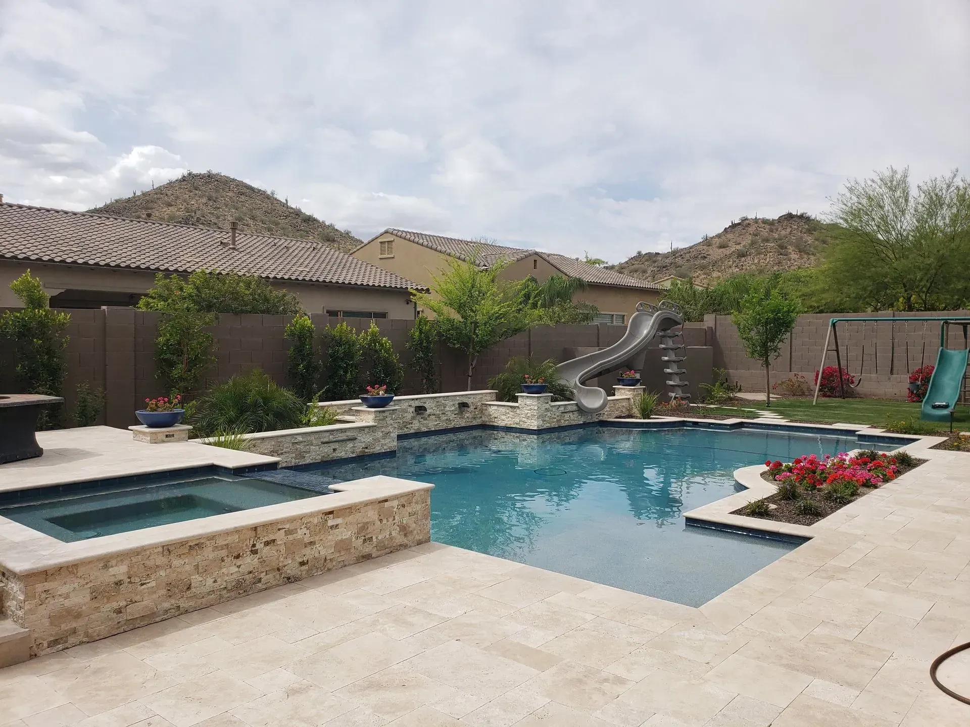 Backyard pool with spa, stone deck, flower bed, and desert-style homes in the background