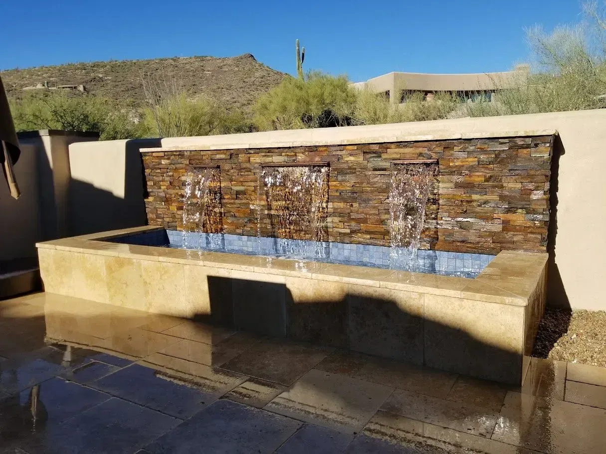 Outdoor stone water fountain with a wooden back wall and desert landscaping behind it