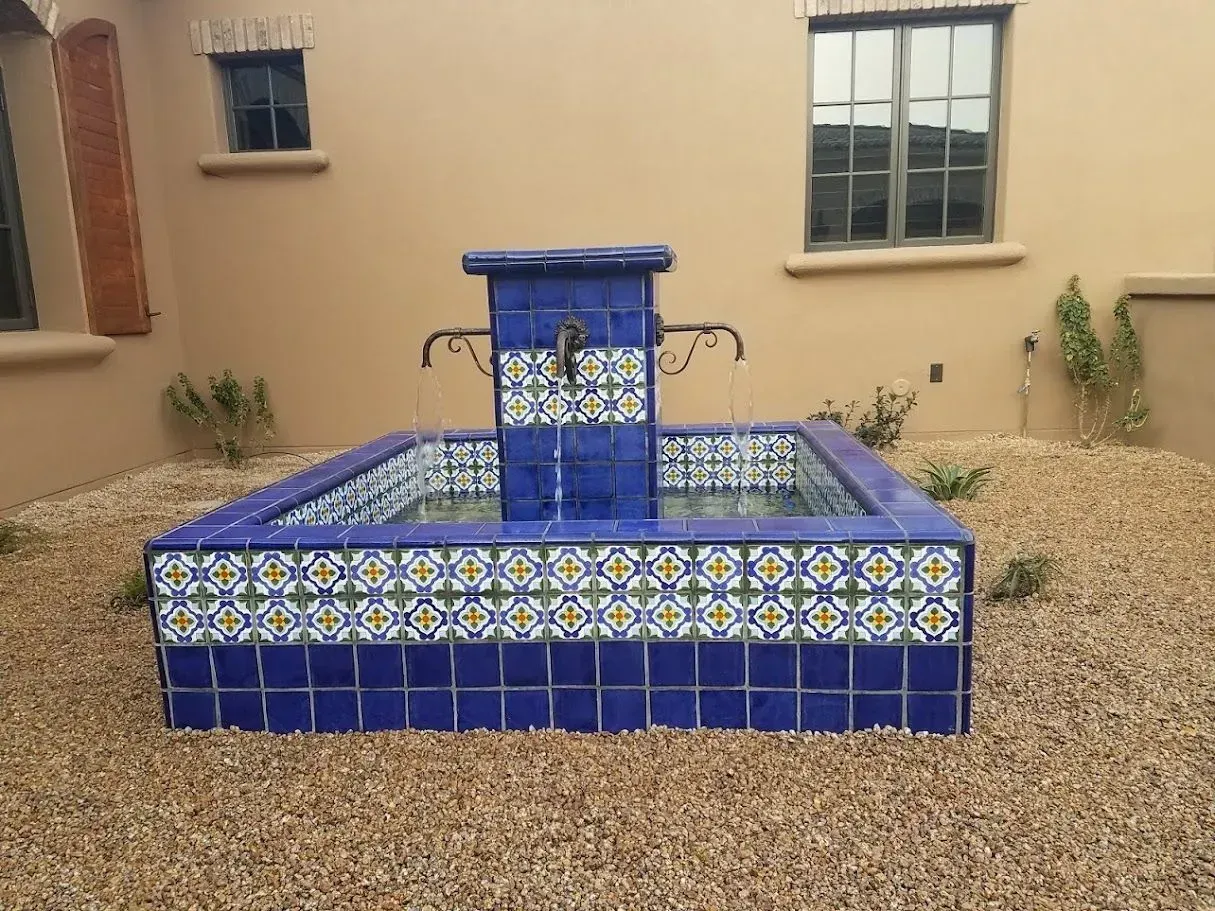 Blue tiled fountain in a gravel courtyard beside a beige building with windows.