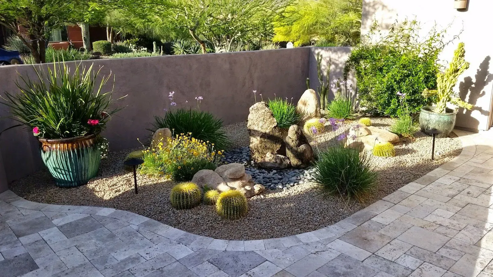 Backyard rock garden with potted plants, shrubs, and decorative stones beside a paved patio