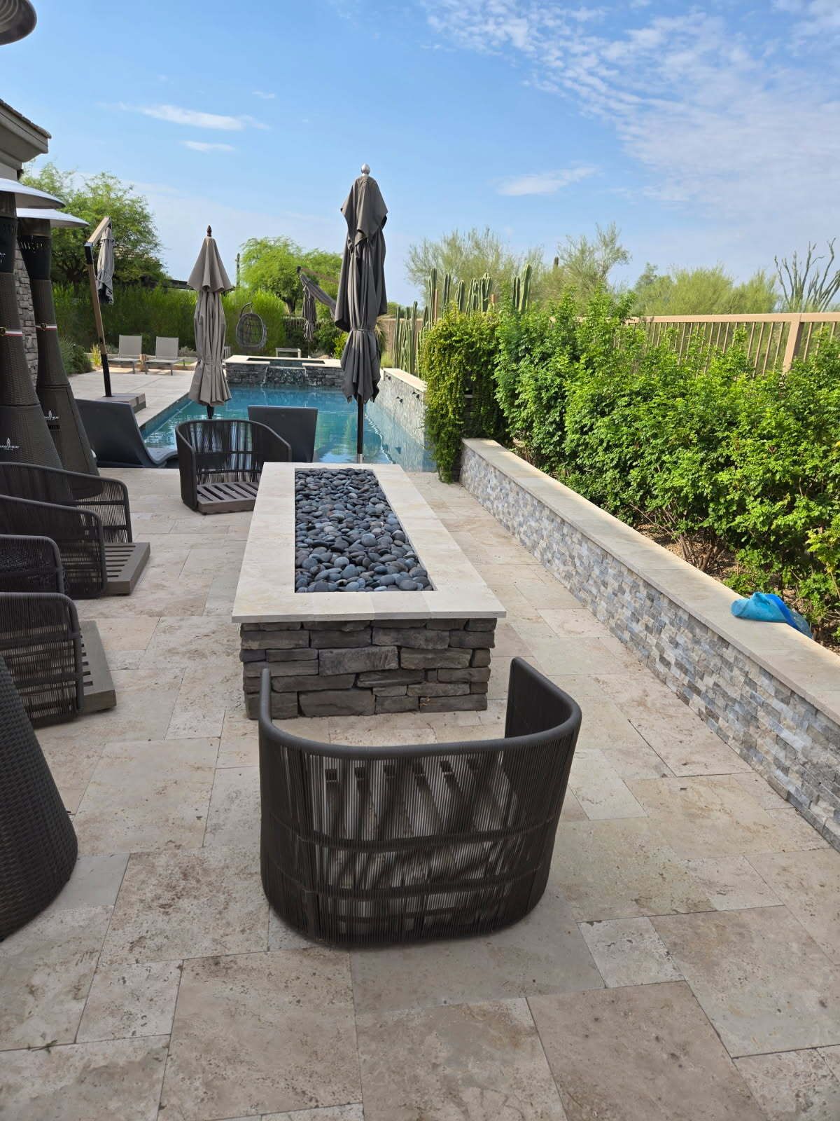 Modern backyard patio with stone fire table, pool, lounge chairs, and tall hedges