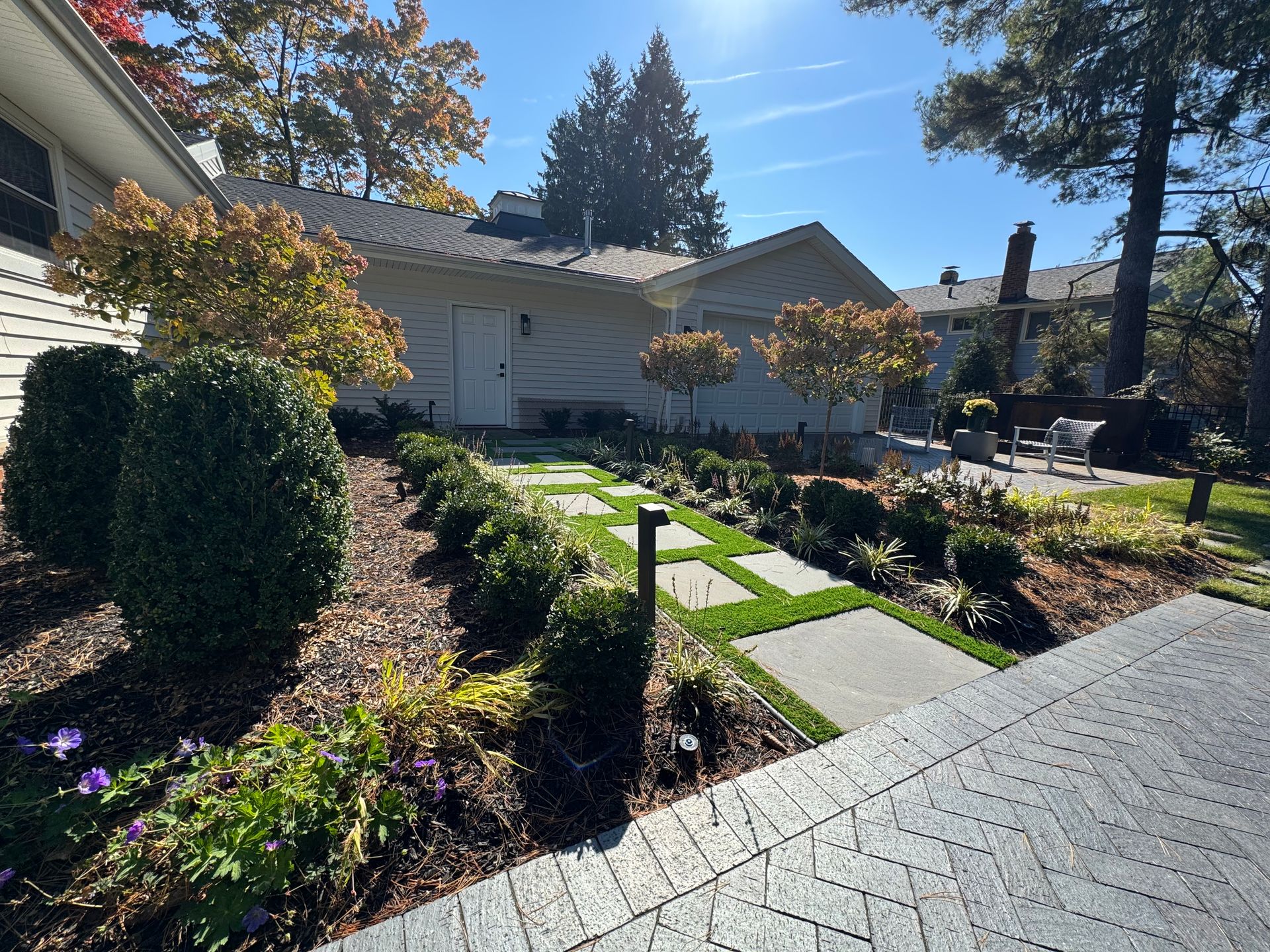 Front yard of a house with a pathway of rectangular stones surrounded by manicured bushes and trees under a clear blue sky on a sunny day.
