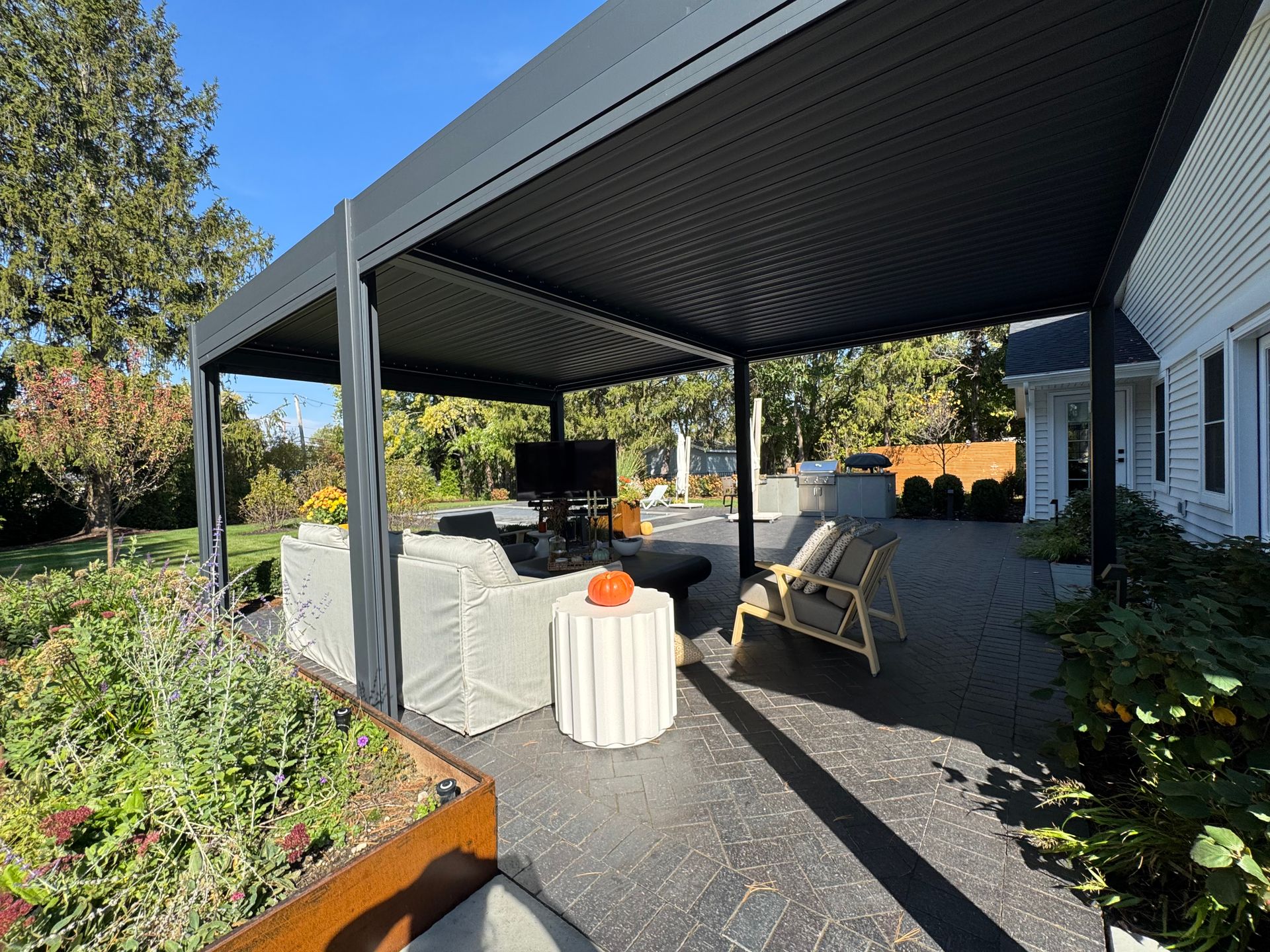 Outdoor patio with a modern pergola featuring sleek gray couches and a central TV. A small table holds an orange pumpkin, adding a cozy touch.