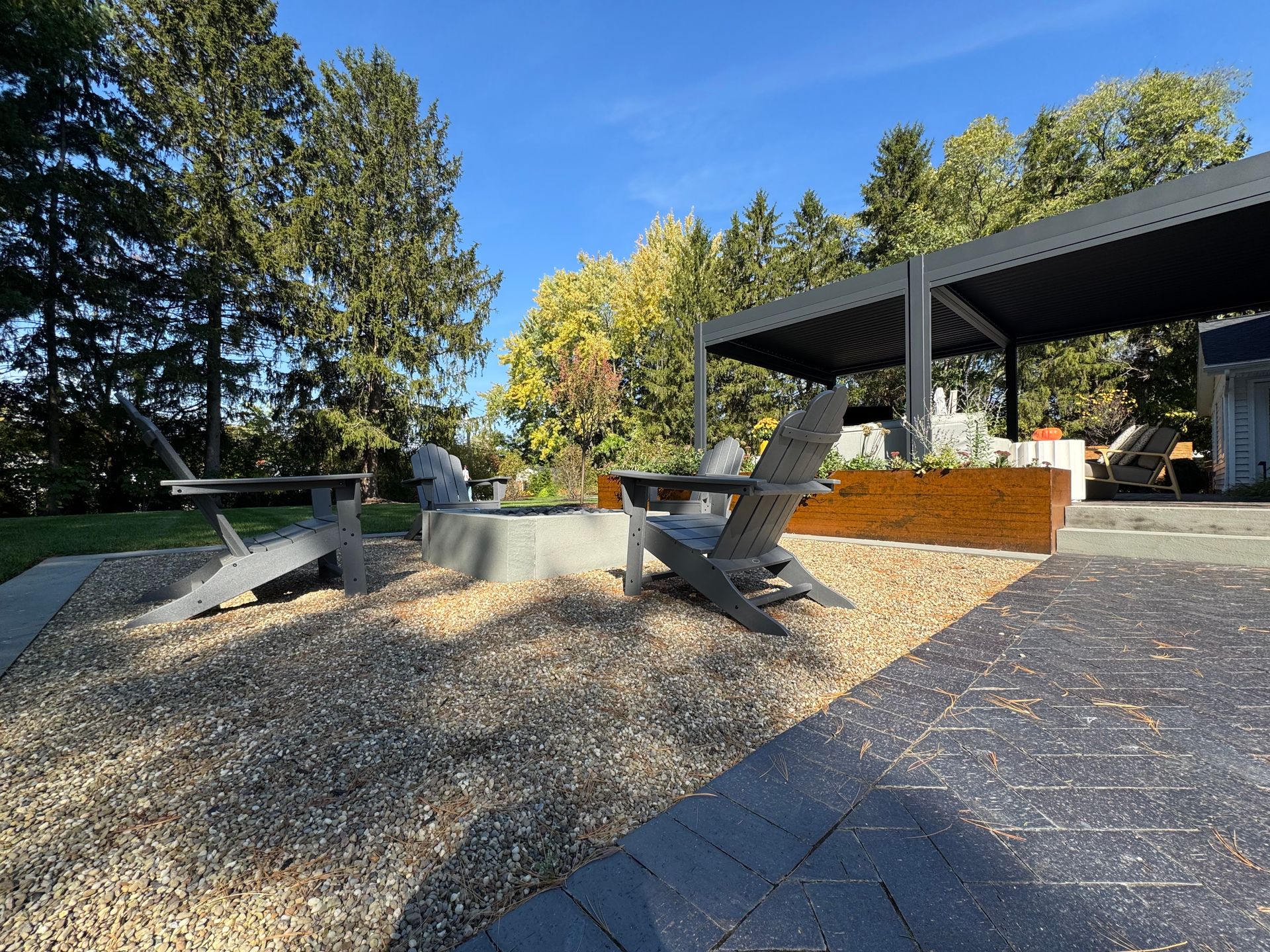 Outdoor patio with Adirondack chairs on gravel surrounding a fire pit. A modern pergola with seating is nearby. Conveys a relaxing, sunny atmosphere.